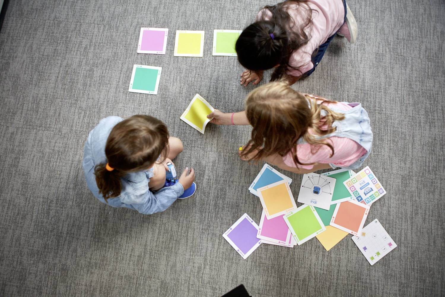 Drei Kinder sitzen auf einem Teppich und spielen mit bunten Karten. Sie scheinen eine Aktivität oder ein Spiel zu veranstalten.