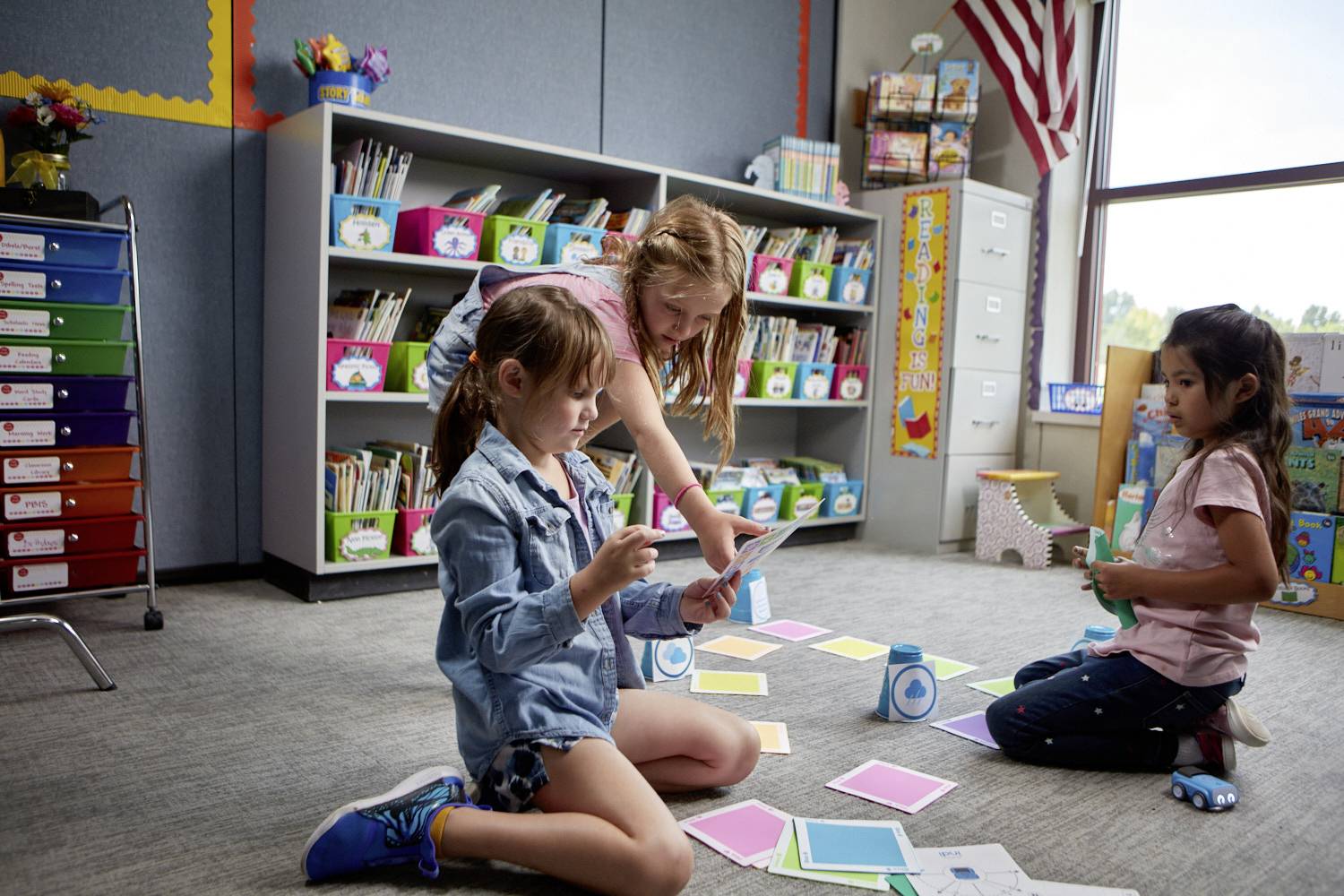 Drei Kinder sitzen auf dem Boden eines Klassenzimmers und arbeiten mit bunten Karten. Im Hintergrund sind Bücherregale und Fenster.
