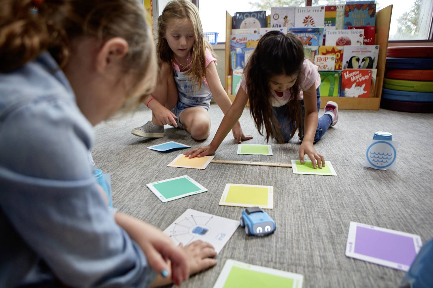 Drei Kinder spielen auf dem Boden mit bunten Karten und einem blauen Spielzeugauto. Im Hintergrund sind Bücher auf Regalen zu sehen.