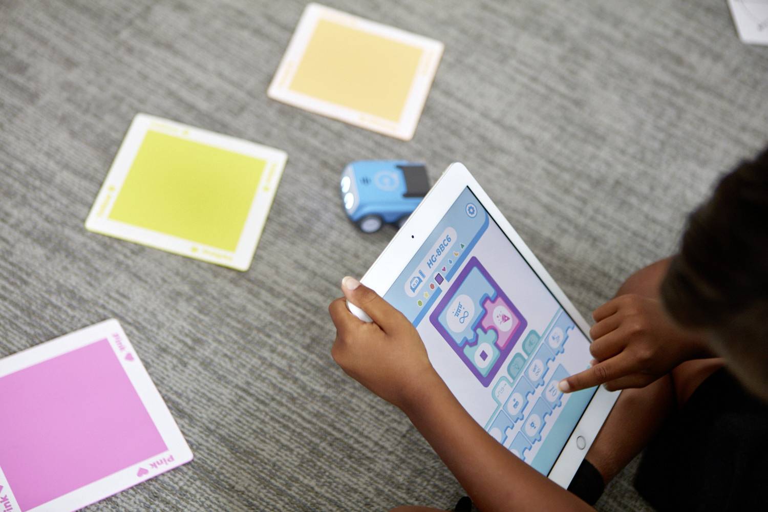 Ein Kind spielt mit einem Tablet, auf dem ein Lernspiel zur Programmierung angezeigt wird, umgeben von bunten Karten und einem Spielzeugroboter.