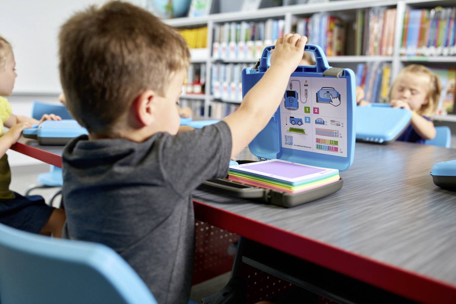 Ein Kind sitzt an einem Tisch in einem Klassenzimmer und benutzt ein Tablet mit Lehrmaterial. Andere Kinder sind im Hintergrund zu sehen.