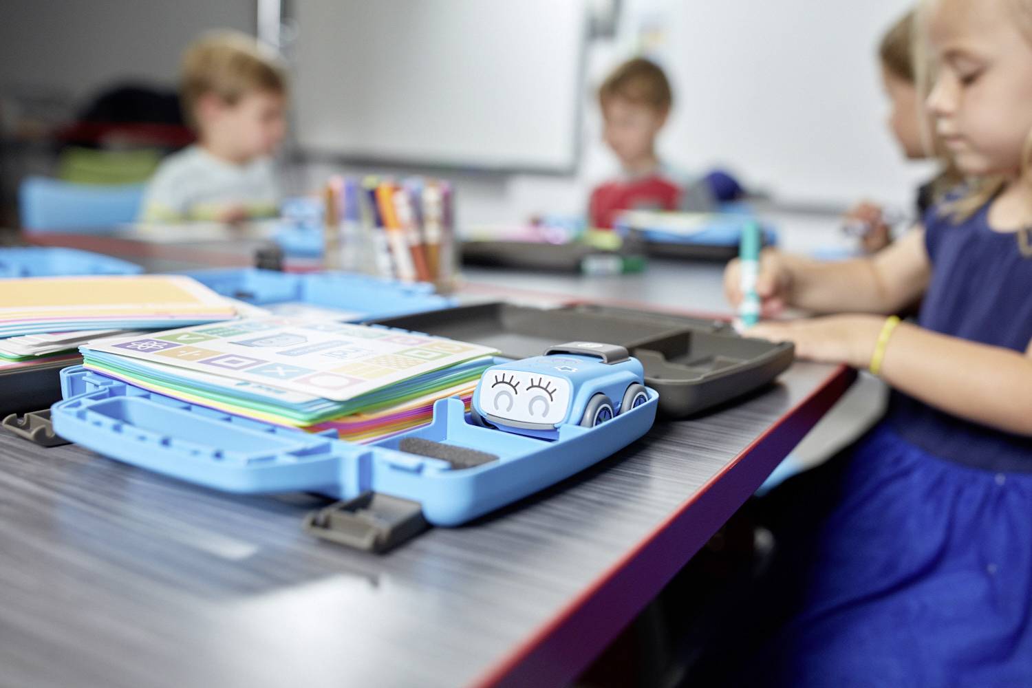 Eine Gruppe von Kindern sitzt in einem Klassenzimmer. Im Vordergrund liegt ein blaues Kunststoff-Kit mit Bastelmaterialien auf dem Tisch.
