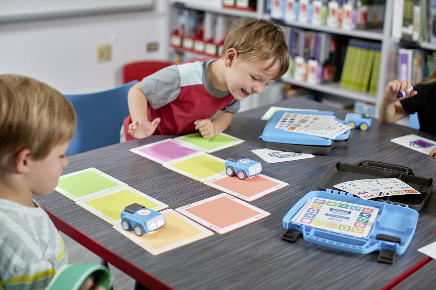 Zwei Kinder spielen mit programmierbaren Robotern auf einem Tisch in einer Bibliothek. Sie sind von Büchern und Materialien umgeben.