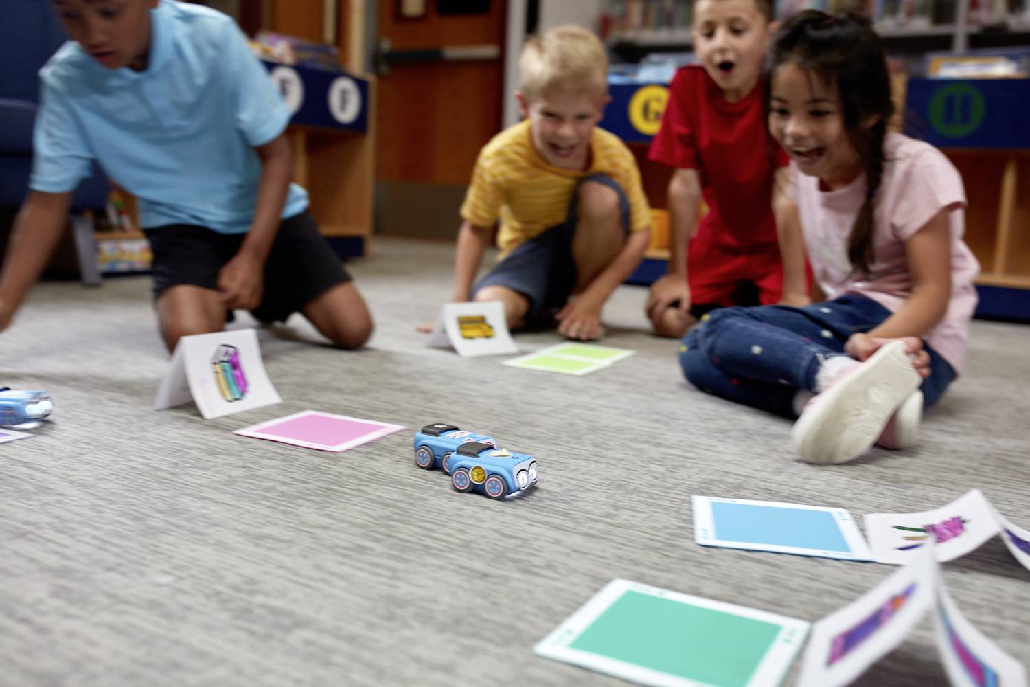 Kinder spielen begeistert auf dem Boden mit kleinen, farbigen Spielzeugautos, die über Karten fahren, in einem hellen Klassenzimmer.
