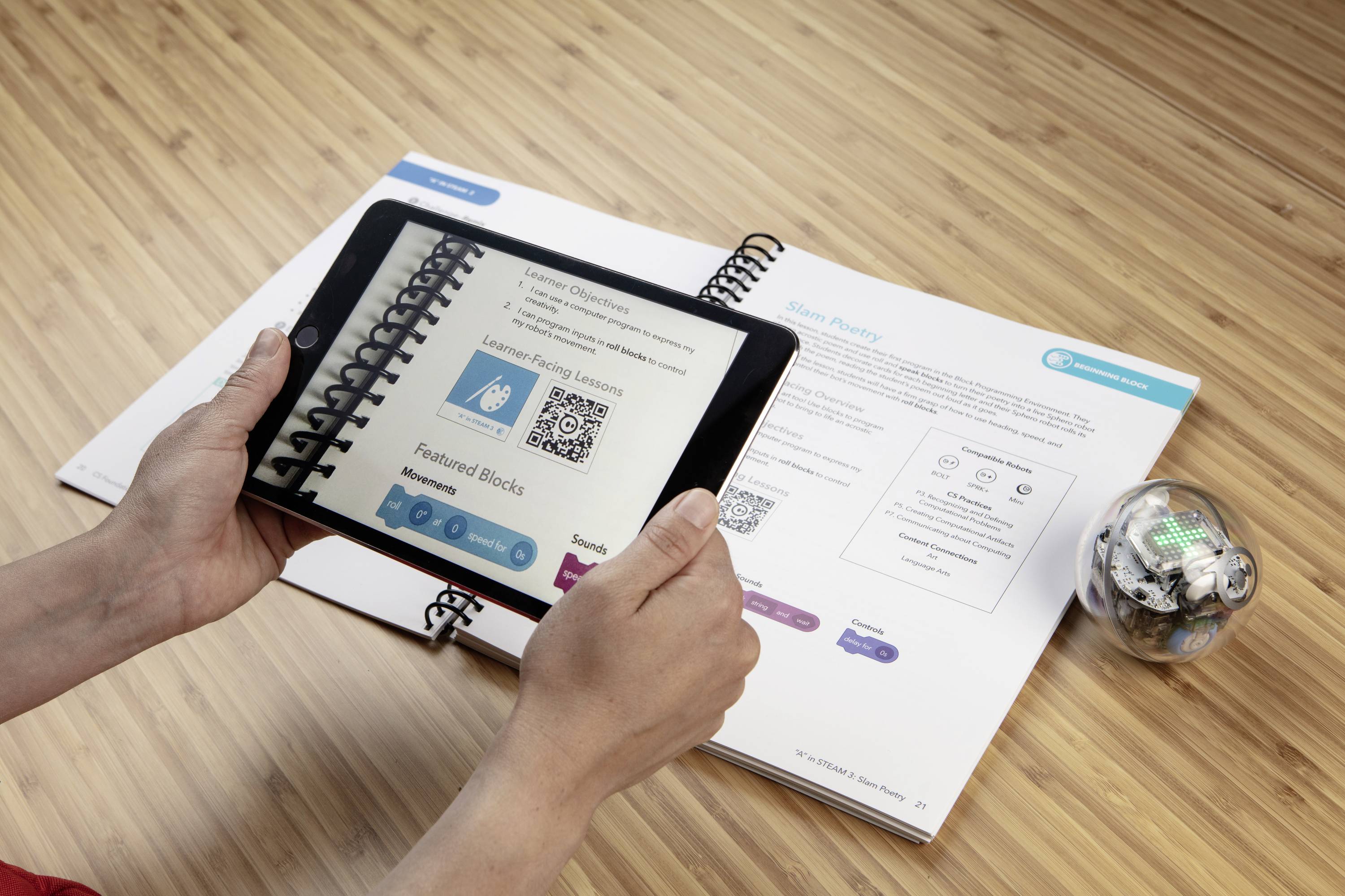 Eine Person hält ein Tablet über ein aufgeschlagenes Buch, um QR-Codes zu scannen. Ein Lernroboter ist rechts neben dem Buch platziert.