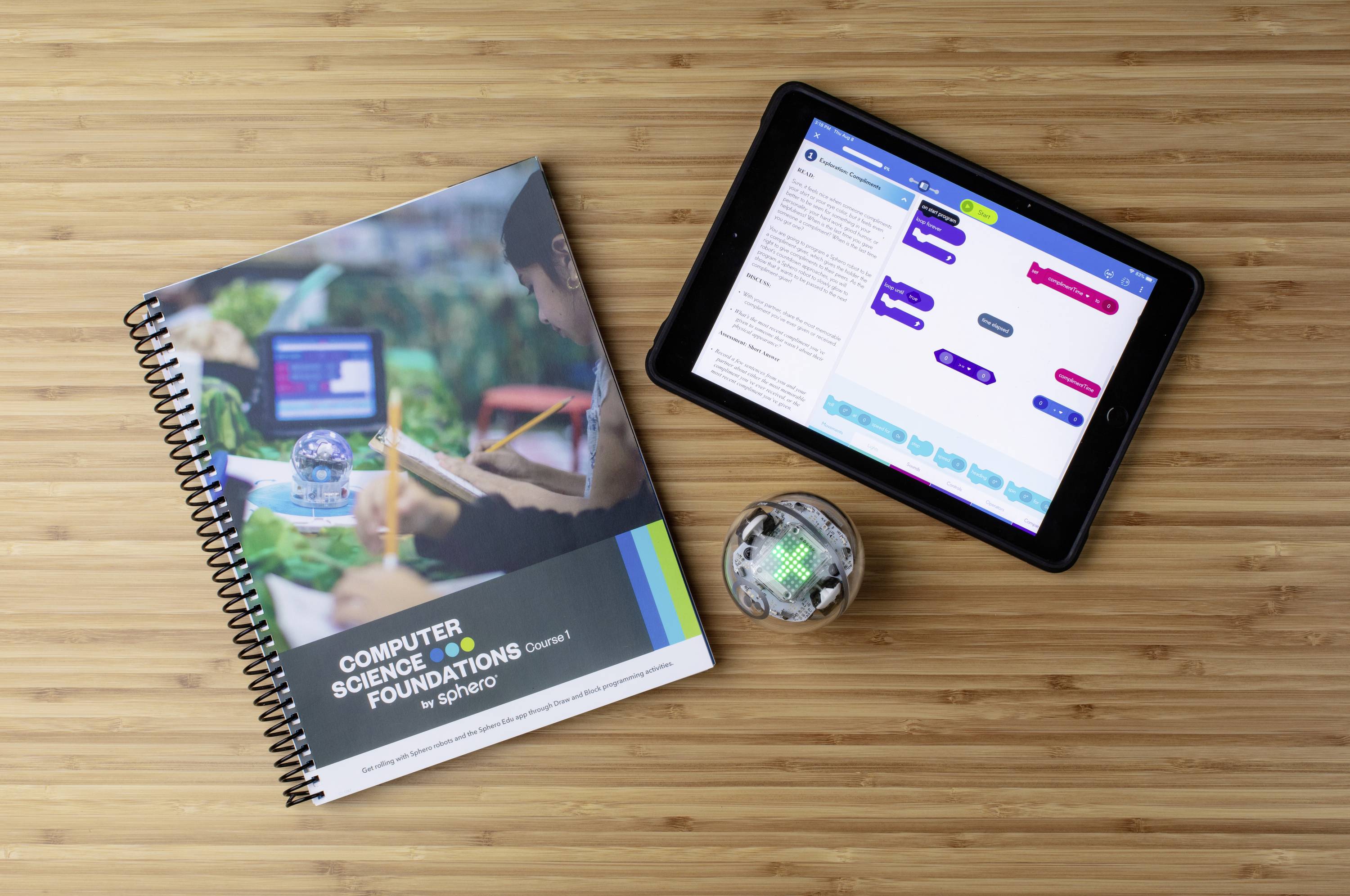 Ein Tablet zeigt eine Programmier-App mit Blockly-Codes; daneben ein Heft 'Computer Science Foundations' und ein kleiner Roboter auf einem Tisch.