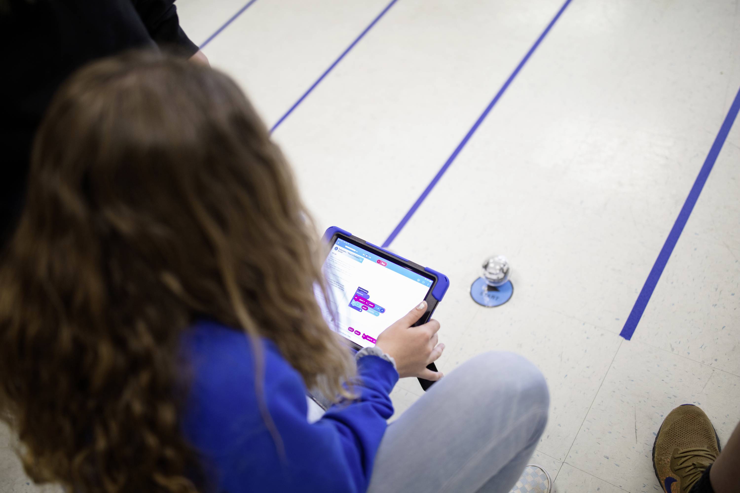 Eine Person steuert mit einem Tablet einen kleinen Roboter auf dem Boden. Der Fokus liegt auf Technologie und Interaktion.