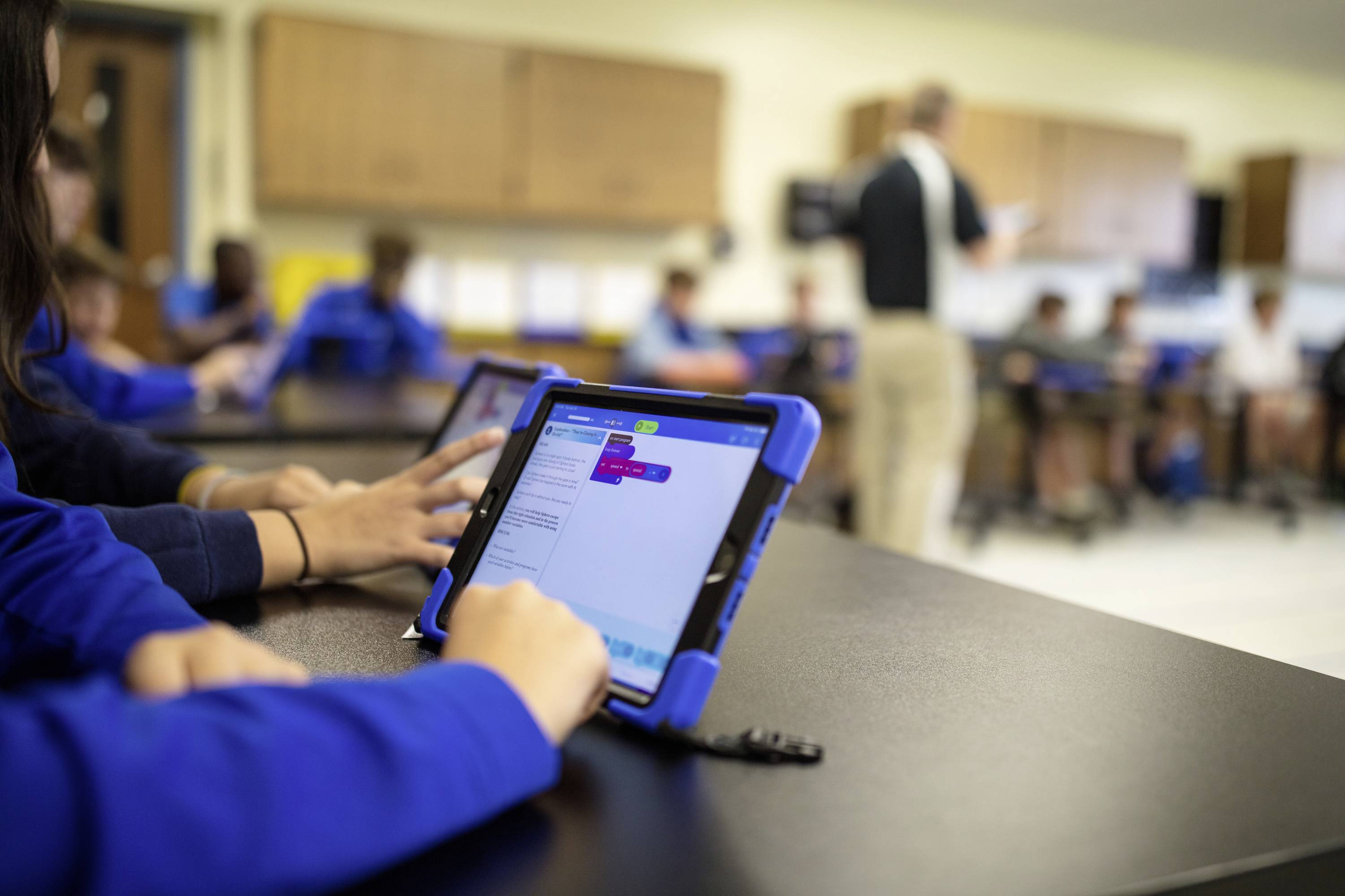 Schüler in einem Klassenraum arbeiten konzentriert auf Tablets an Programmieraufgaben. Ein Lehrer steht im Hintergrund und unterrichtet.