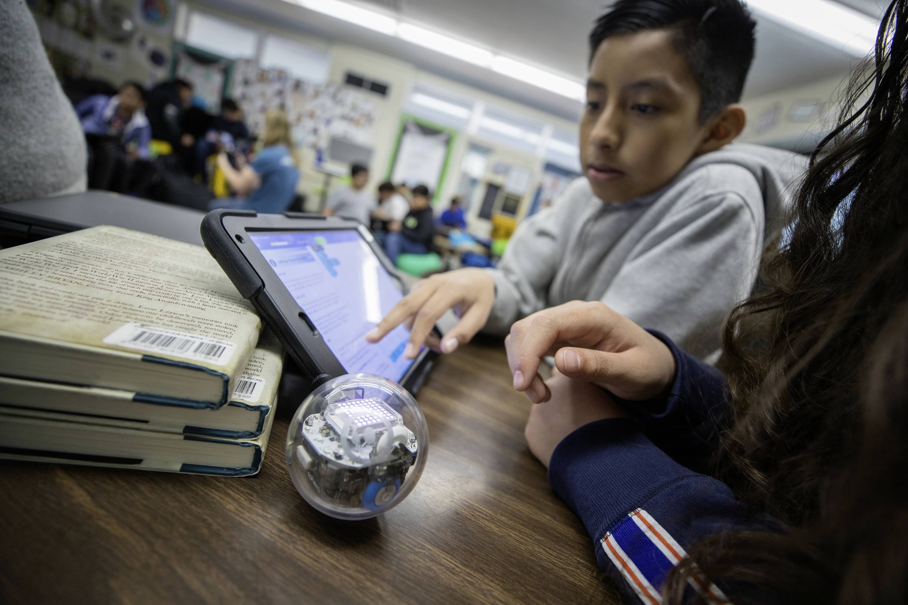 Ein Junge und ein Mädchen programmieren einen kleinen Roboterball mit einem Tablet in einem Klassenzimmer. Bücher liegen daneben.