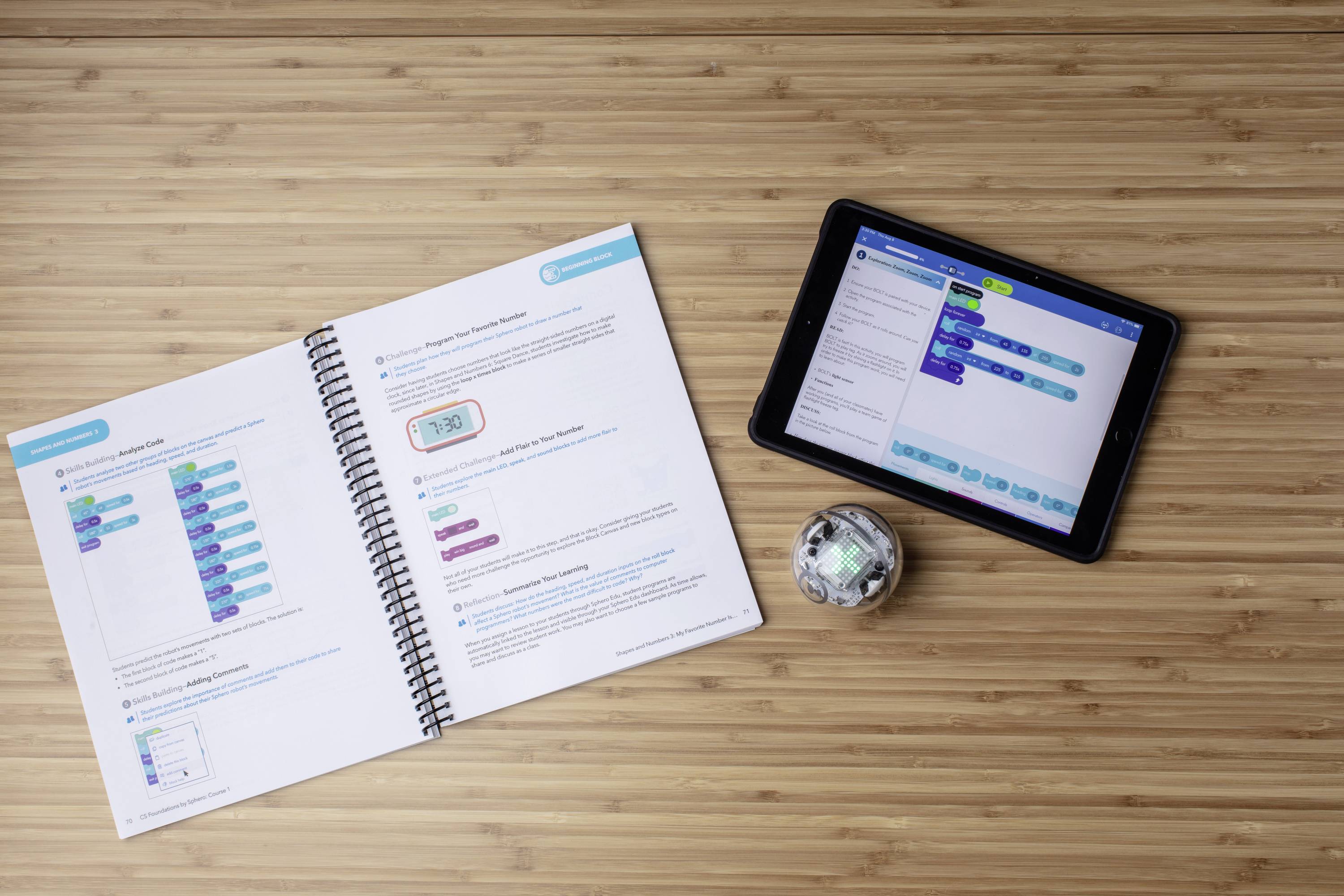 Ein offenes Lehrbuch und ein Tablet mit einer Programmierschnittstelle liegen auf einem Tisch. Daneben ein kleiner Roboterball.