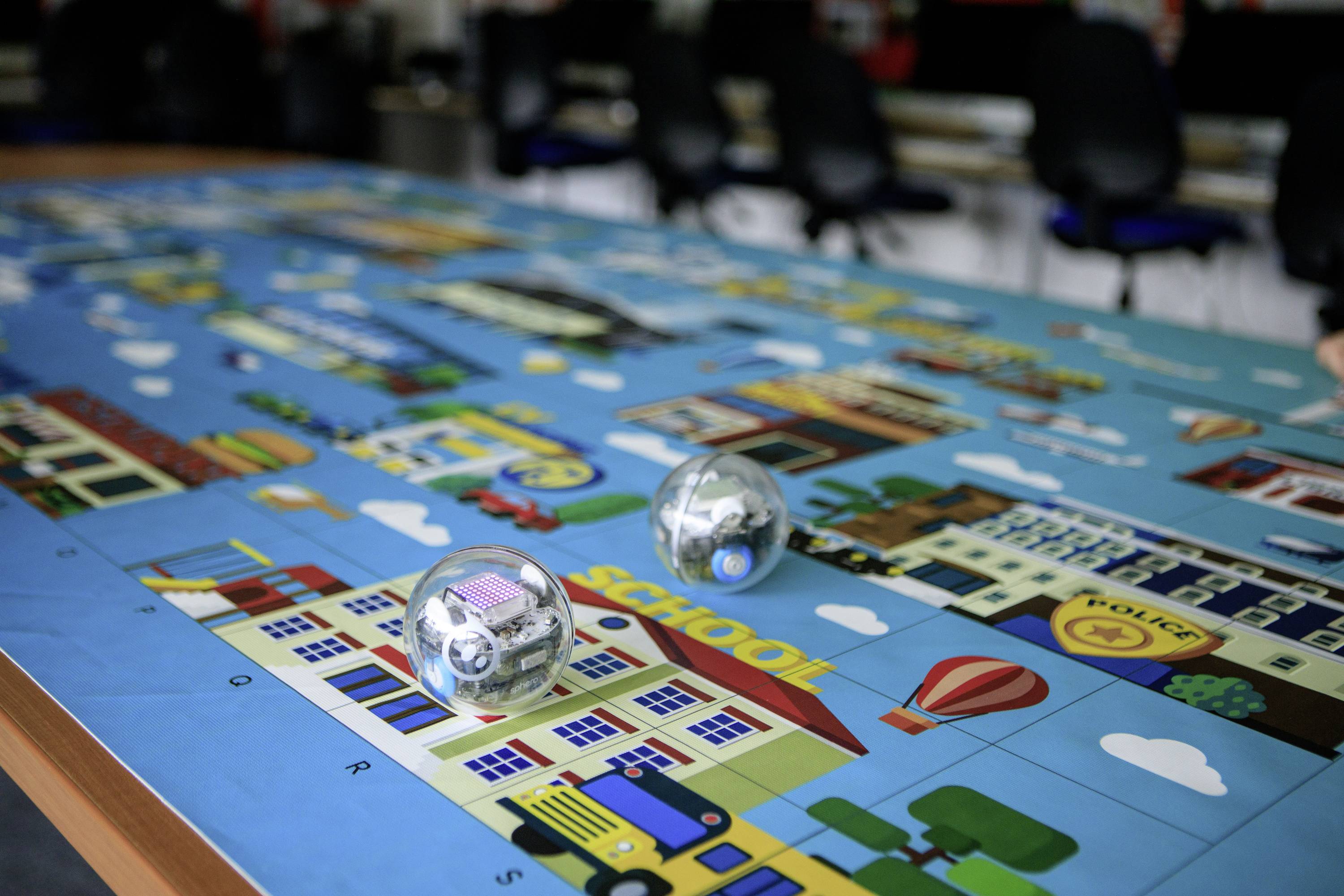 Zwei Roboterkugeln bewegen sich über eine bunte Spielmatte mit verschiedenen Gebäuden und Straßen. Im Hintergrund sind unscharfe Stühle zu sehen.