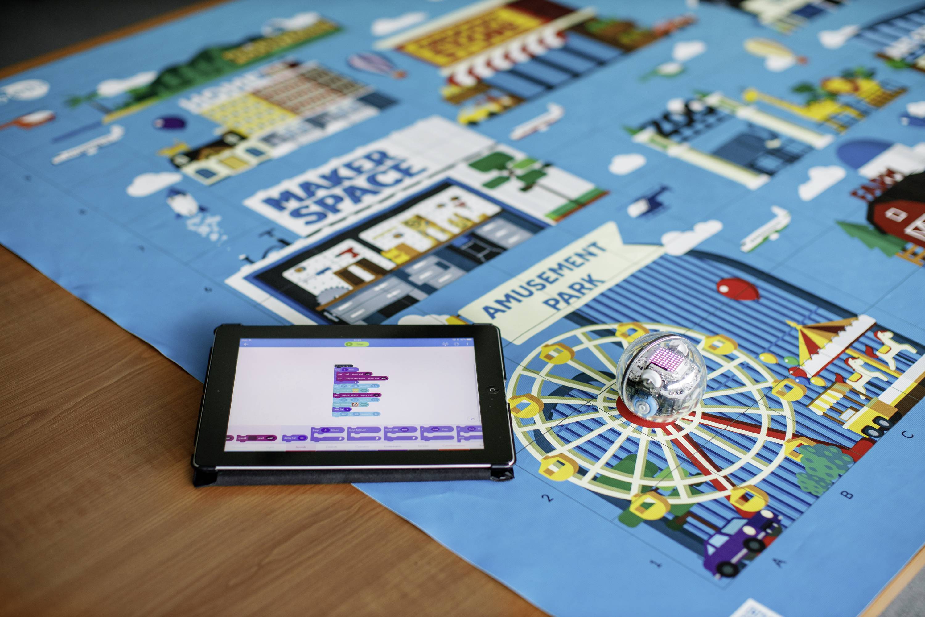 Tablet mit Programmiercode neben einem Roboter auf Spielfeld einer Lernmatte mit Beschriftungen 'Maker Space' und 'Amusement Park'.