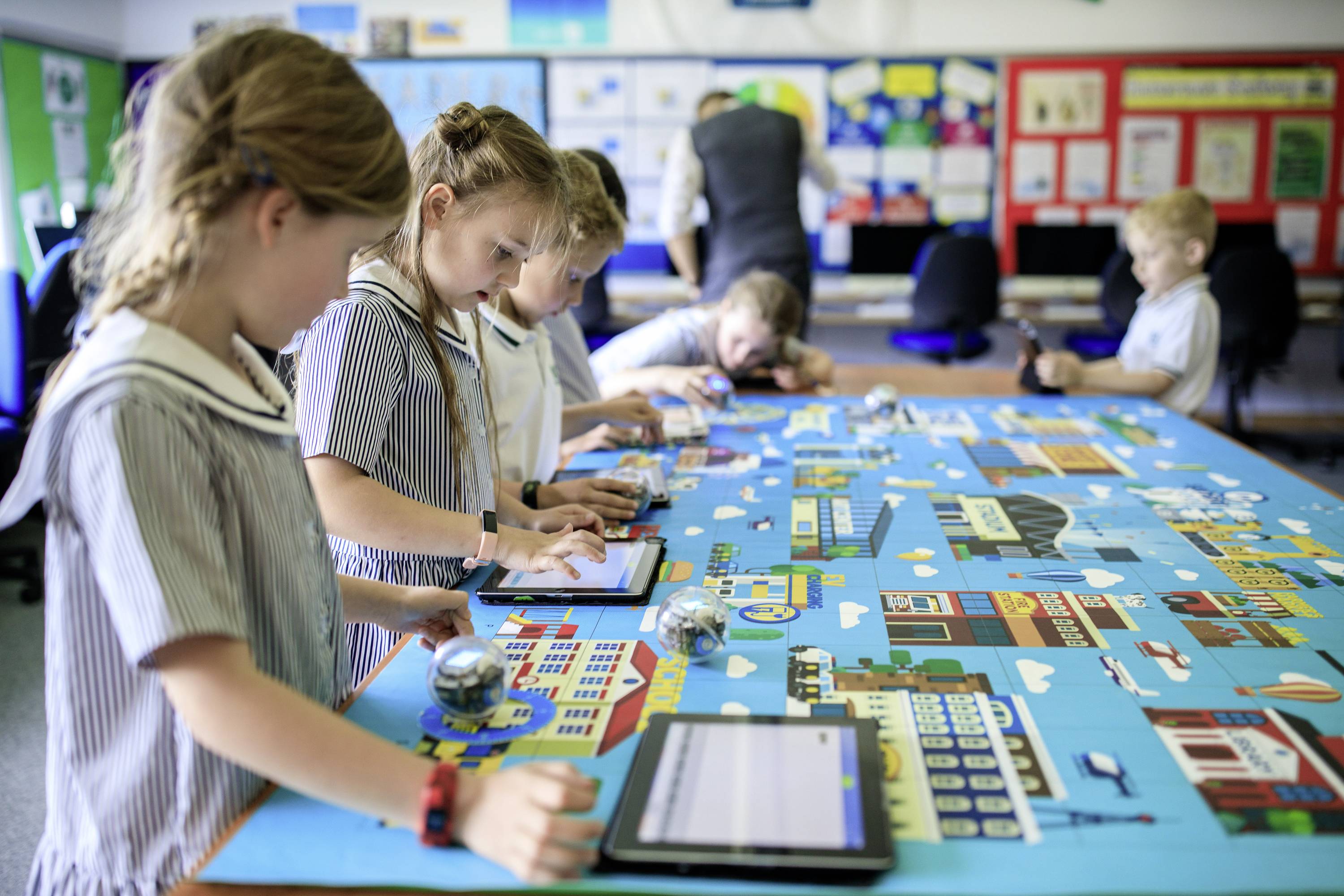 Kinder in Schuluniformen benutzen Tablets und kleine Roboter auf einem großen Tisch mit einem Stadtplan. Im Hintergrund sind Computer sichtbar.