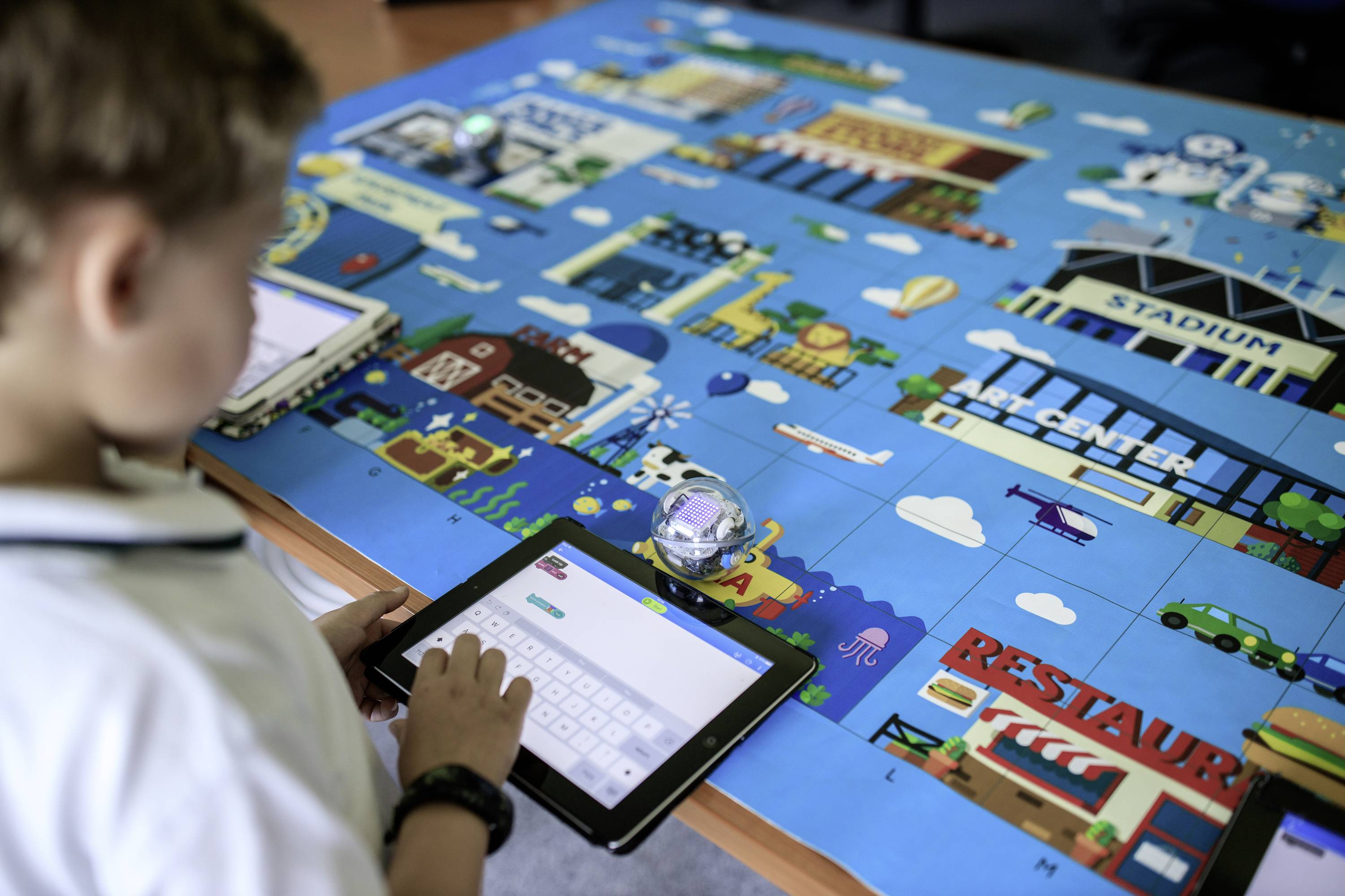 Ein Kind nutzt ein Tablet, um einen Roboter über eine bunte, spielartige Stadtkarte zu steuern. In der Umgebung sind Gebäude und Symbole sichtbar.