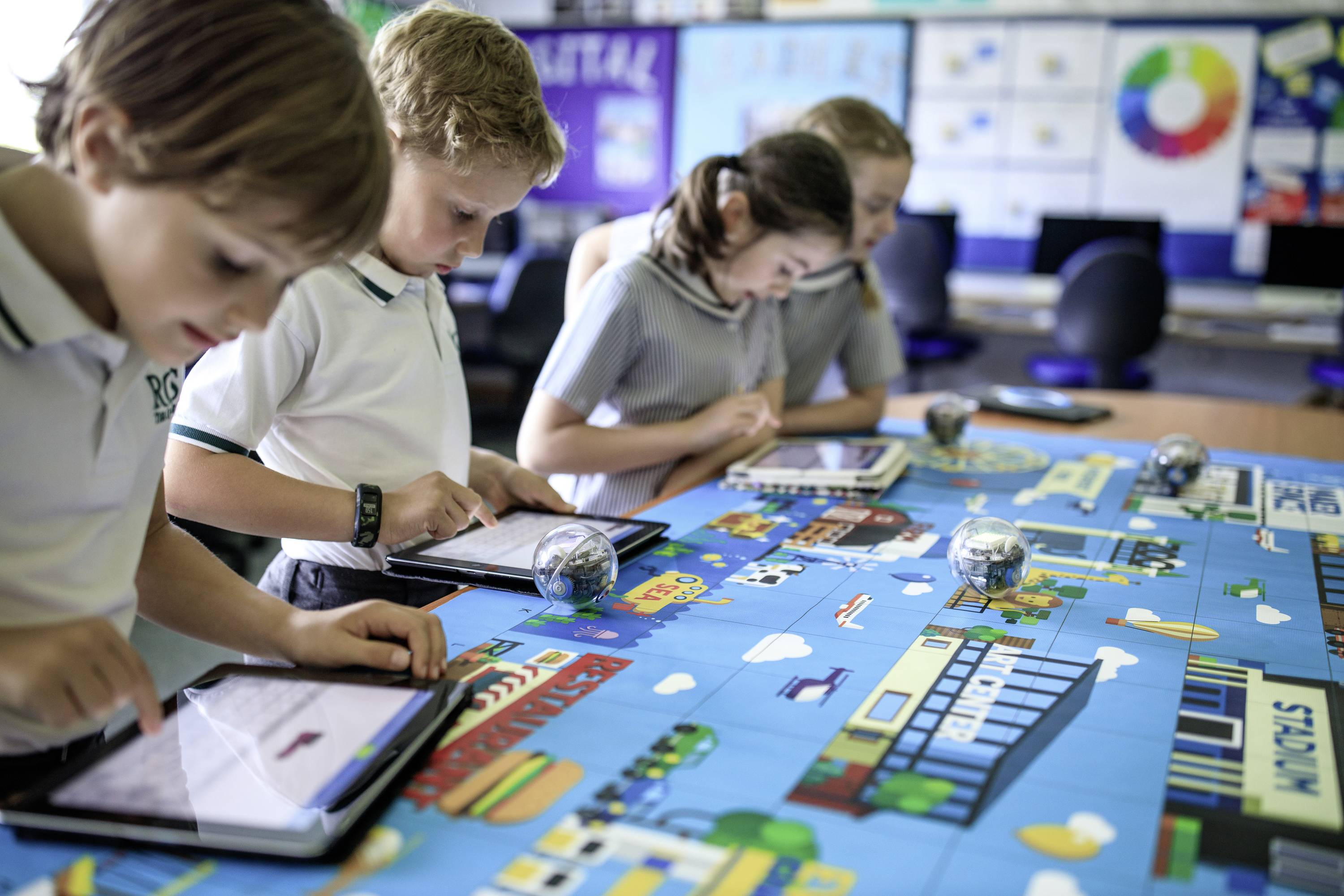 Kinder in Schuluniformen arbeiten konzentriert mit Tablets zusammen an einem bunten Tisch, der mit einem Lernspiel bedeckt ist.