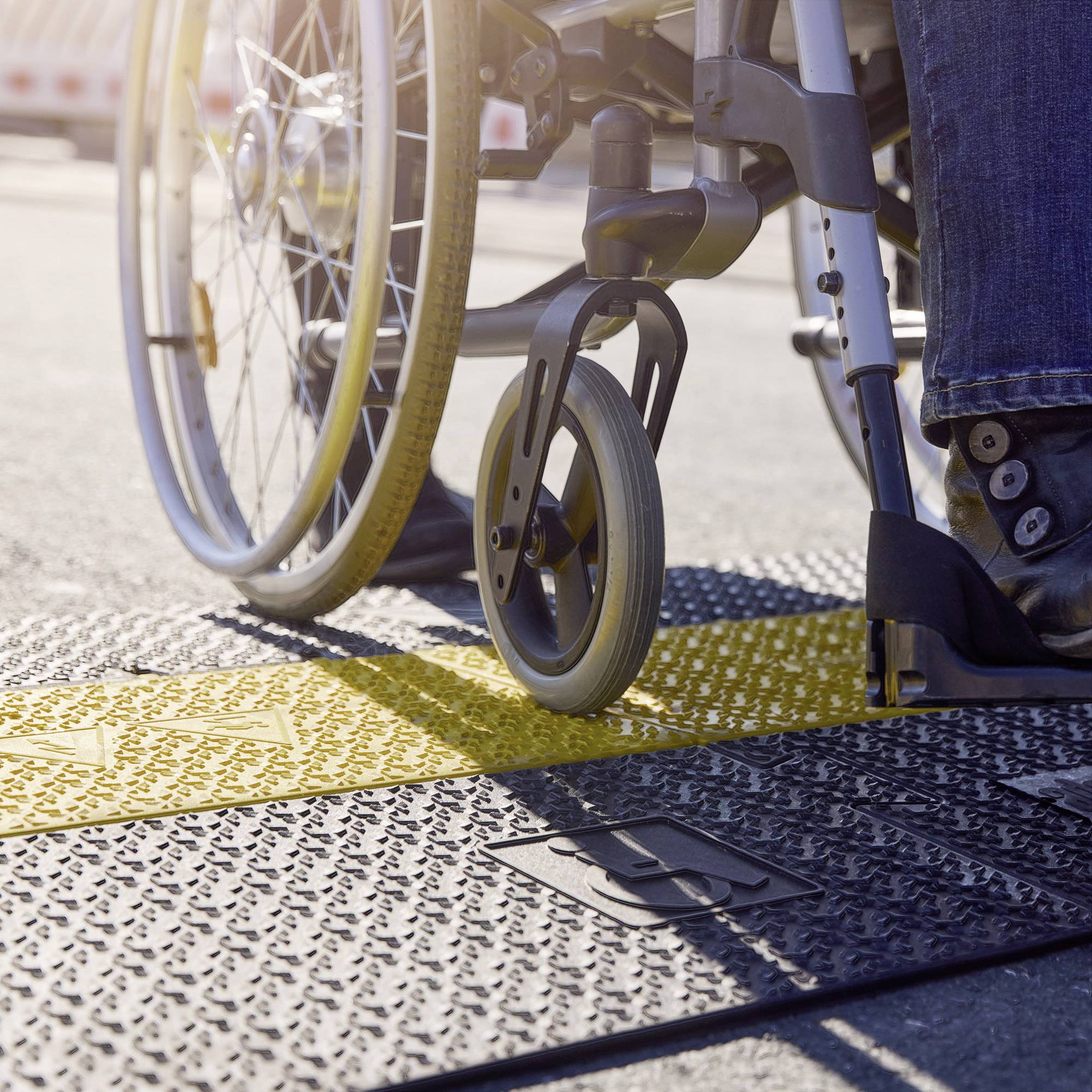 Person im Rollstuhl überquert eine Straße auf einem taktilen Weg. Details der Räder und der strukturierten Bodenplatten sind sichtbar.