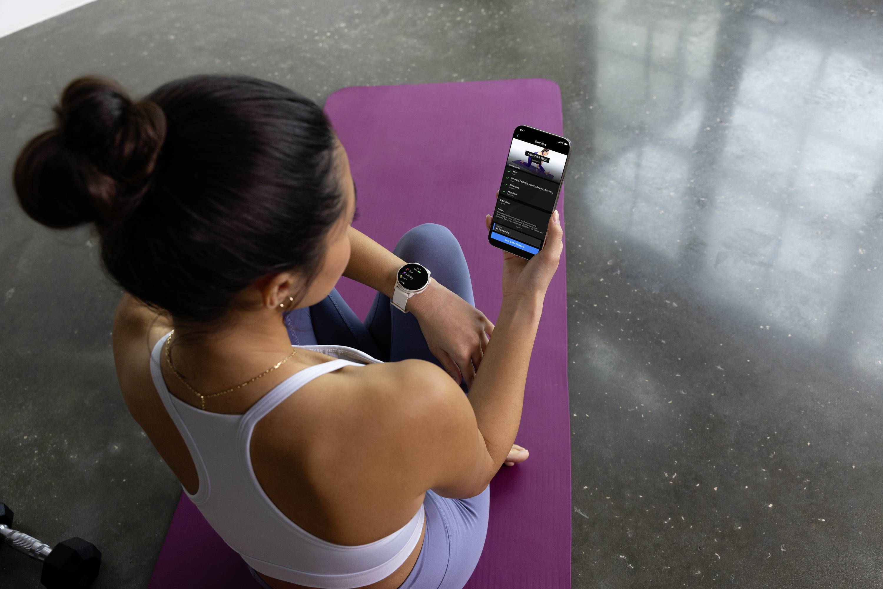 Frau sitzt auf einer lila Yogamatte und schaut auf ihr Smartphone, auf dem eine Fitness-App geöffnet ist. Sie trägt Sportkleidung.