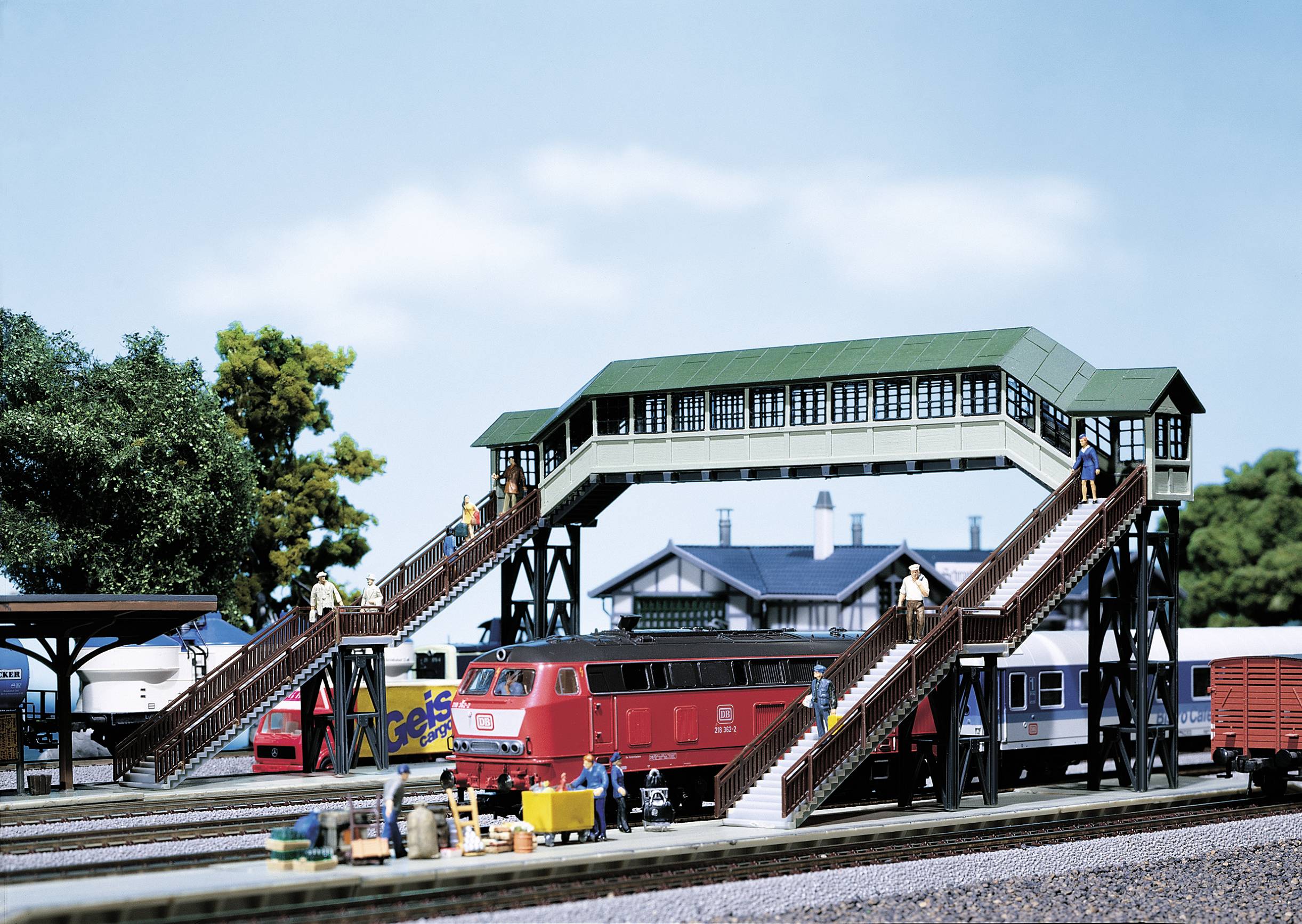 Ein Modellbahnhof mit einer roten Lokomotive auf den Gleisen. Eine Überführung mit grünen Dächern verbindet die Bahnsteige. Im Hintergrund Bäume und ein Gebäude.