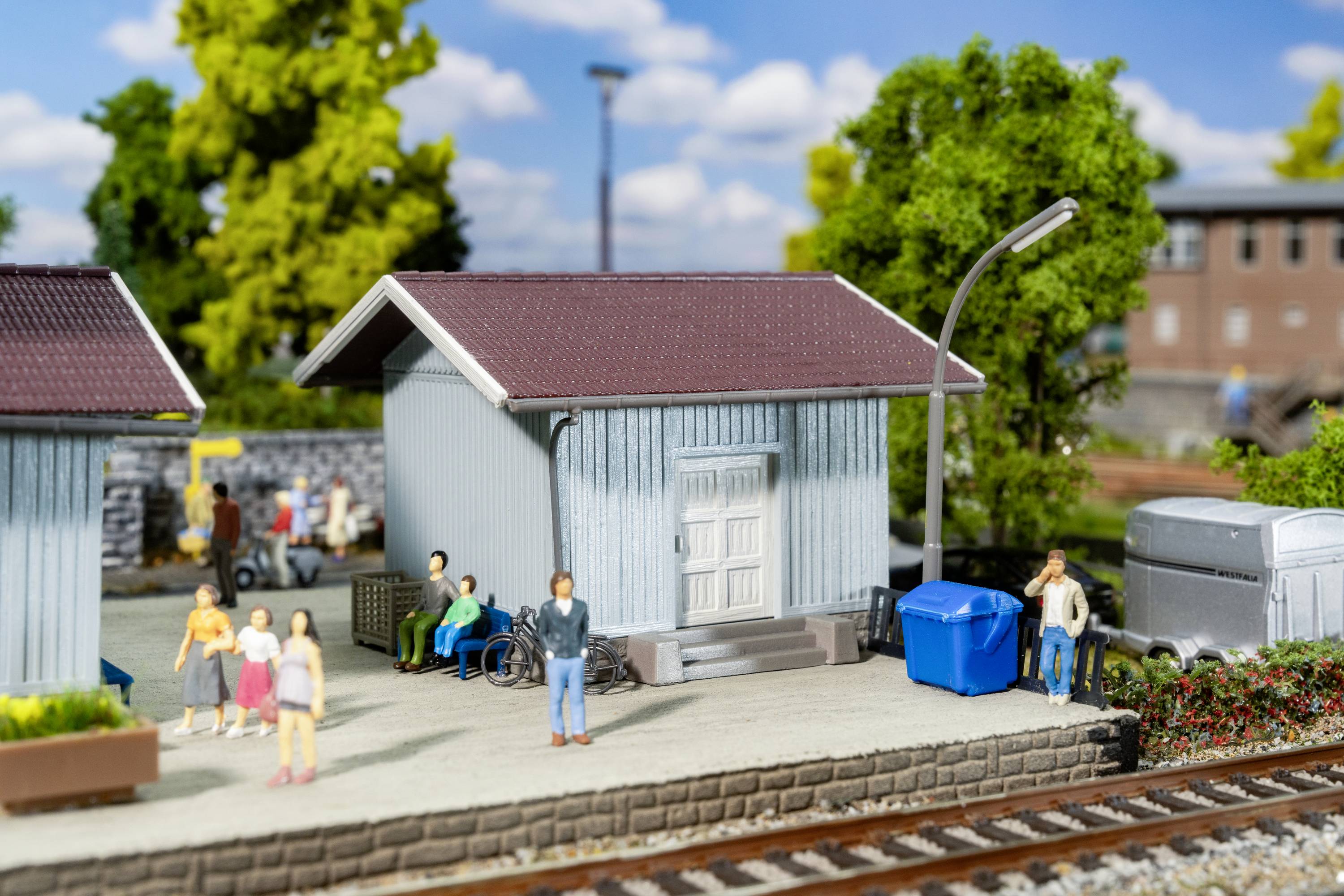 Eine Miniaturszene zeigt eine Bahnhofsumgebung mit kleinen Figuren, darunter Menschen und Rollstuhlfahrer, sowie Gebäude und Bahnübergang.