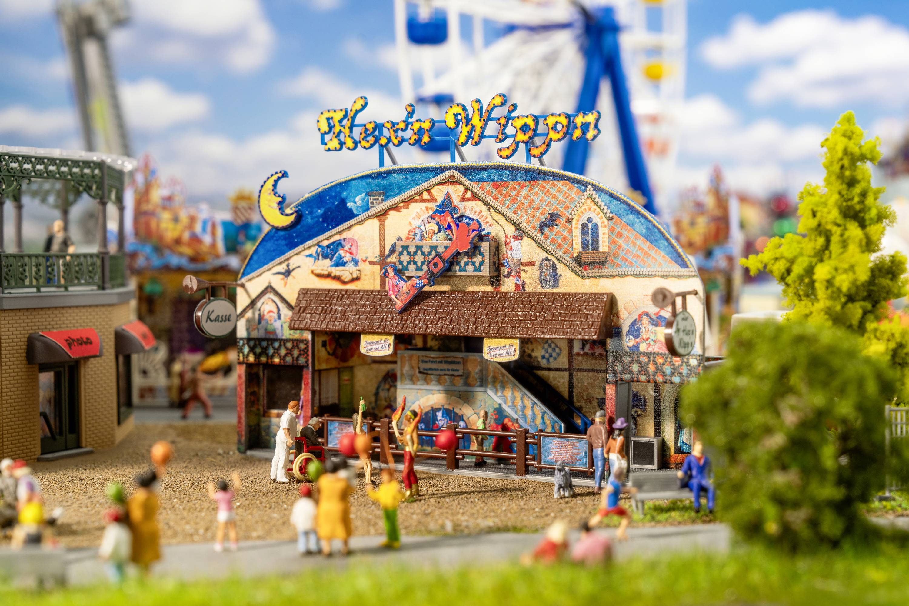 Eine Miniatur-Kirmeszene mit einem bunten 'Hexen Wagen' Zelt, umgeben von Figuren und Bäumen, vor einem blauen Himmel mit Wolken.