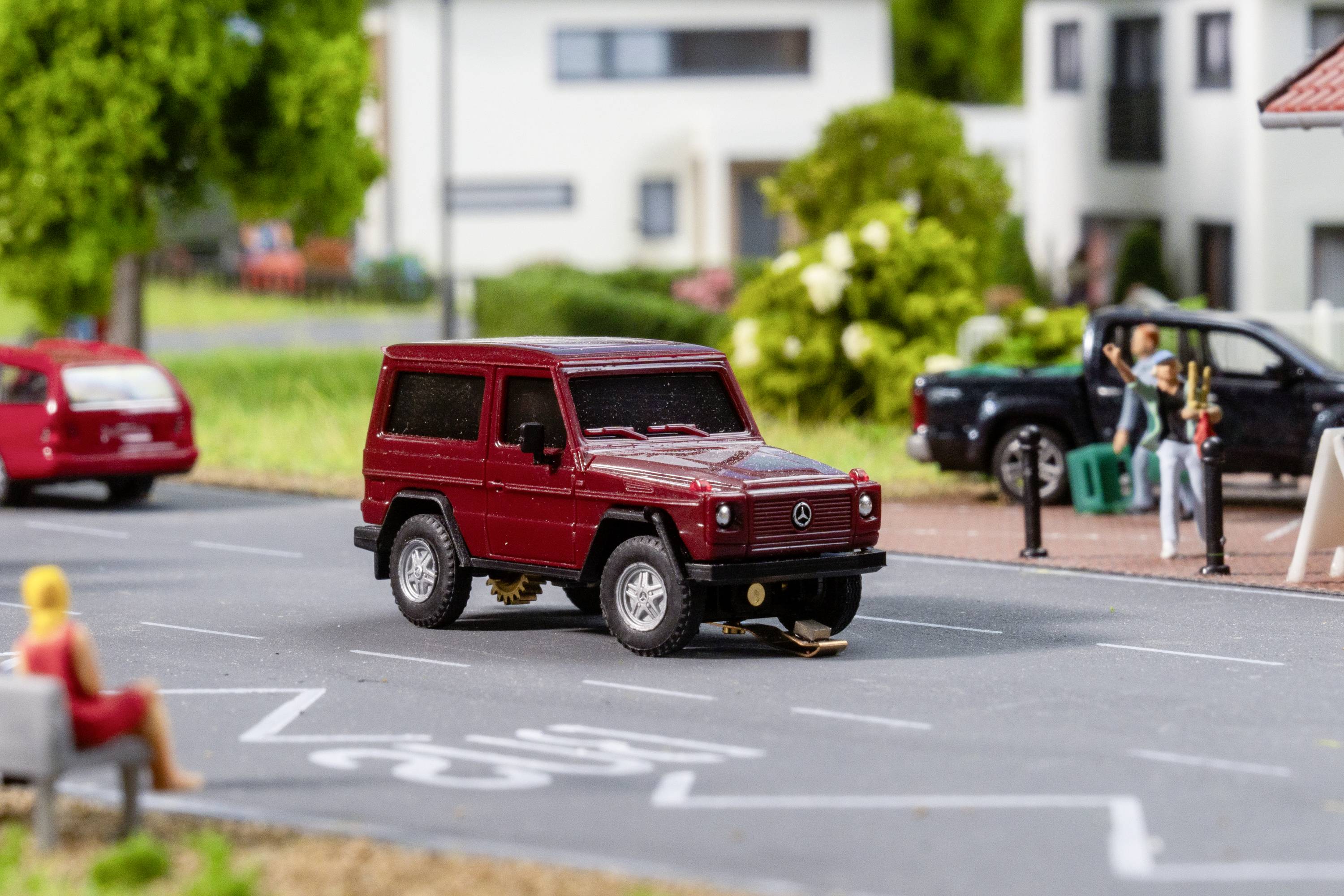 Rotes Modellauto auf Miniaturstraße, umgeben von kleinen Bäumen und Häusern. Im Hintergrund sind Miniaturfiguren sichtbar.
