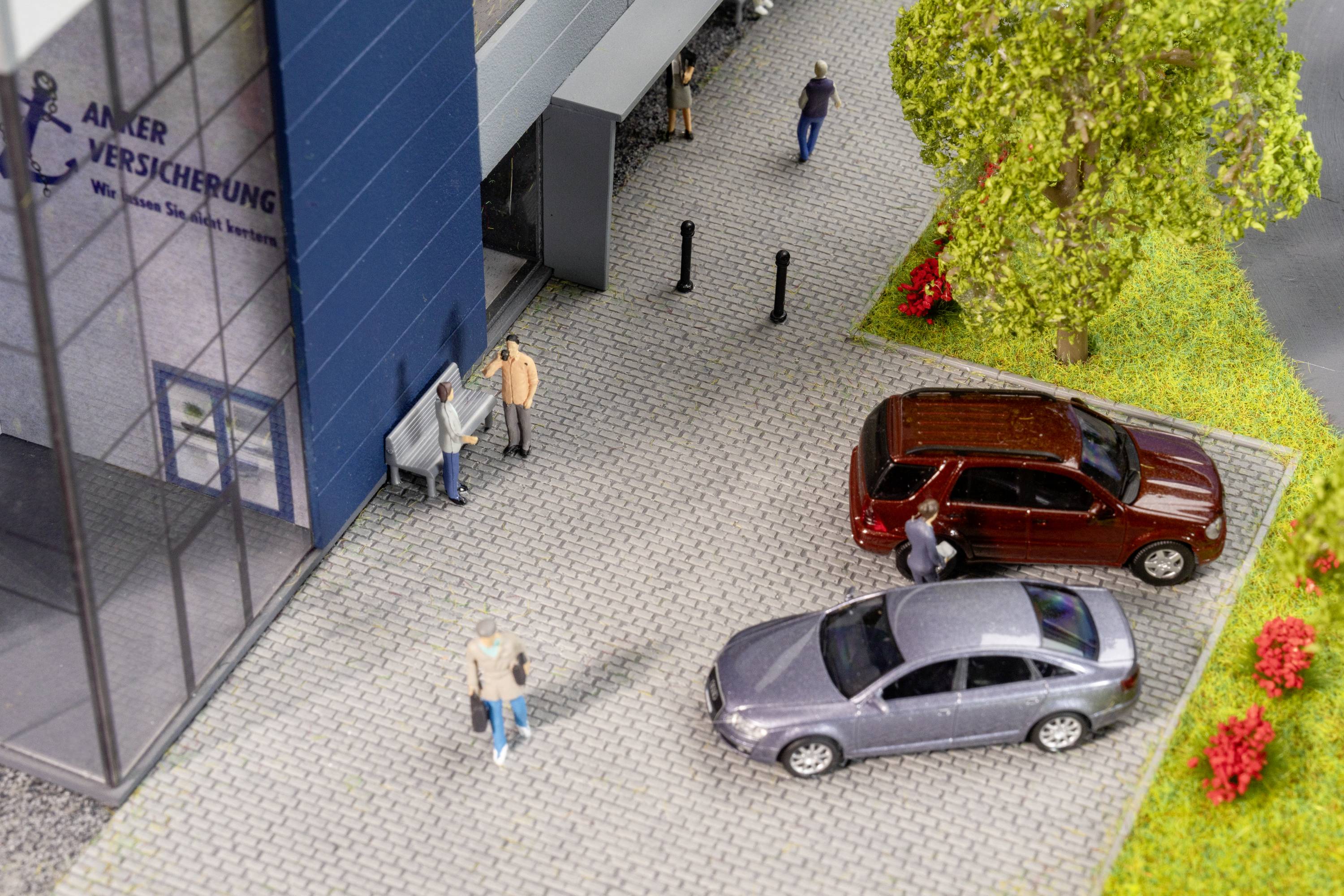 Modell einer Versicherungsagentur. Menschen vor dem Gebäude, zwei geparkte Autos, Bäume und Blumen.