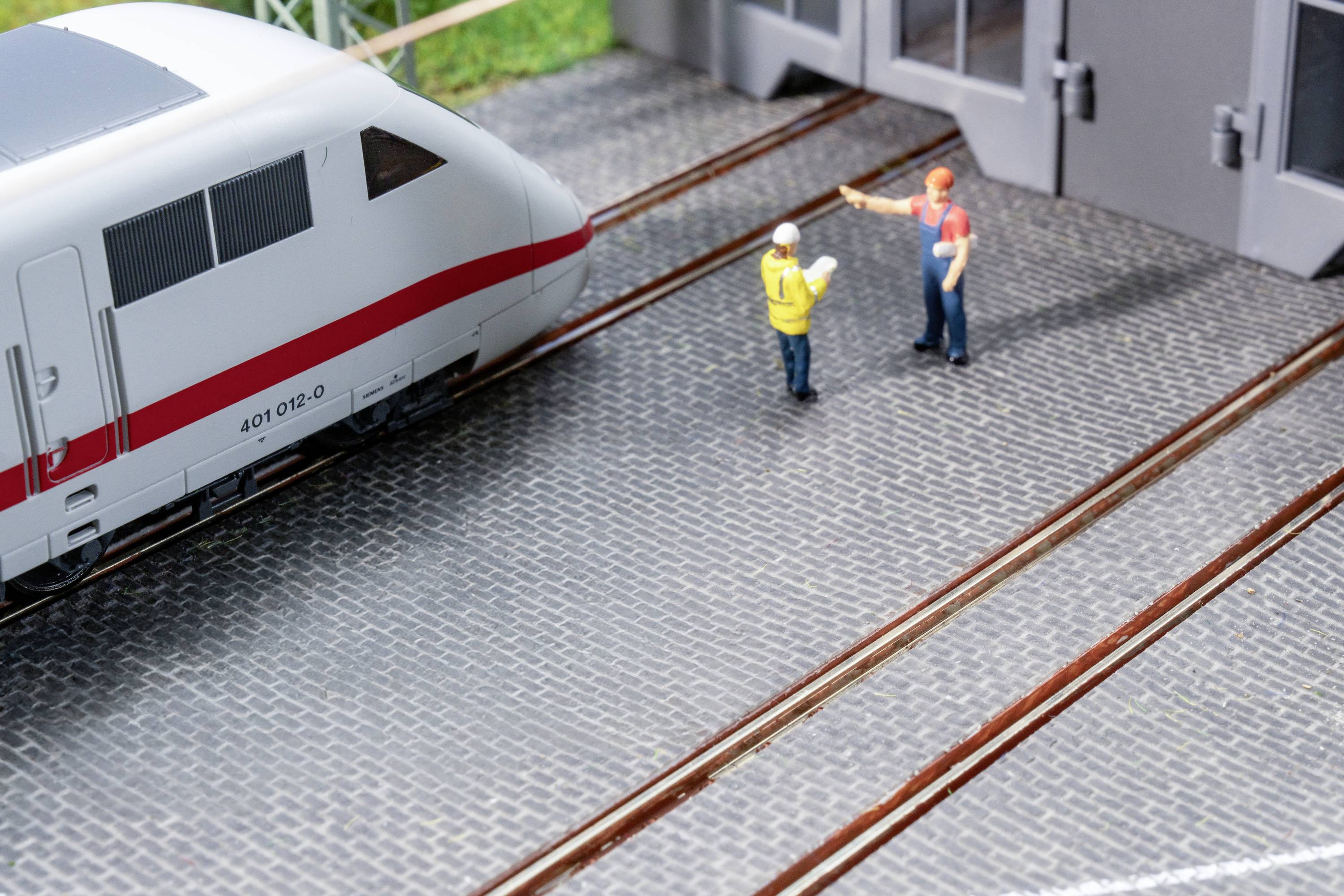 Ein Modellzug hält vor einem Bahnhof. Zwei Figuren in Arbeitskleidung, einer gelb, einer rot, stehen auf den Gleisen und gestikulieren.