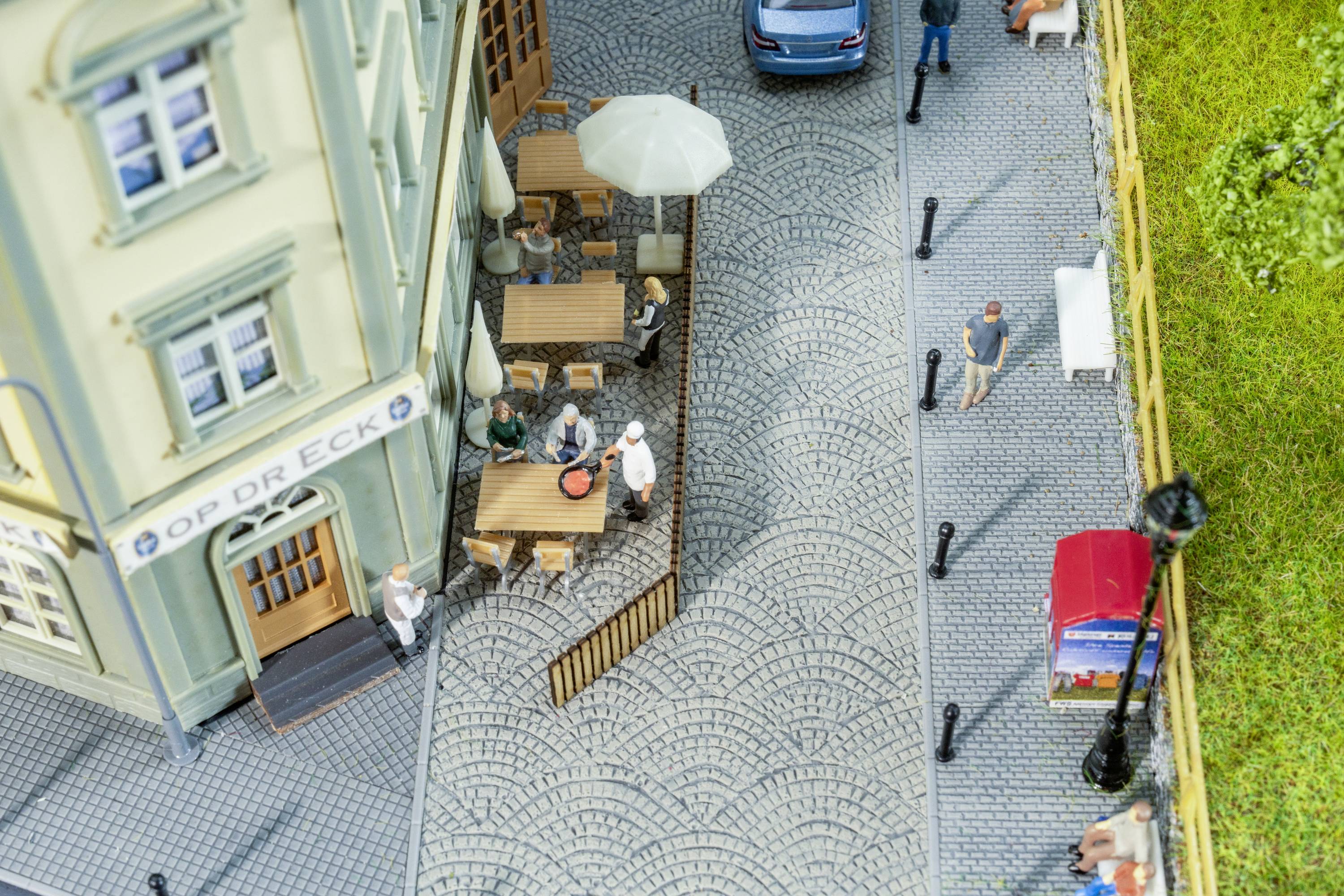 Miniaturmodell von einem Bürgersteig mit Café. Drei Tische, Gäste essen und trinken. Ein blauer Wagen auf der Straße, Menschen spazieren.