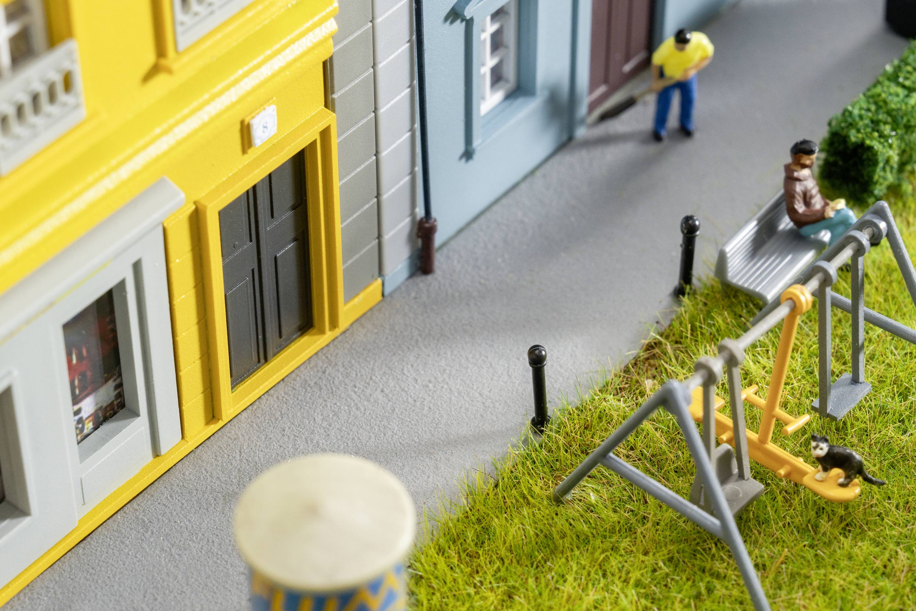 Miniaturmodell einer Stadt mit gelbem Gebäude, Gehweg, Schaukel und Parkbank. Zwei Miniaturfiguren sind zu sehen, eine auf der Bank.
