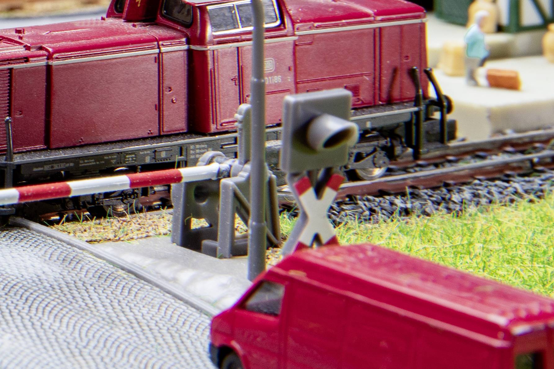 Ein rotes Spielzeugmodell eines Zuges fährt über einen Bahnübergang, mit einer Schranke und Straßensperre im Vordergrund.