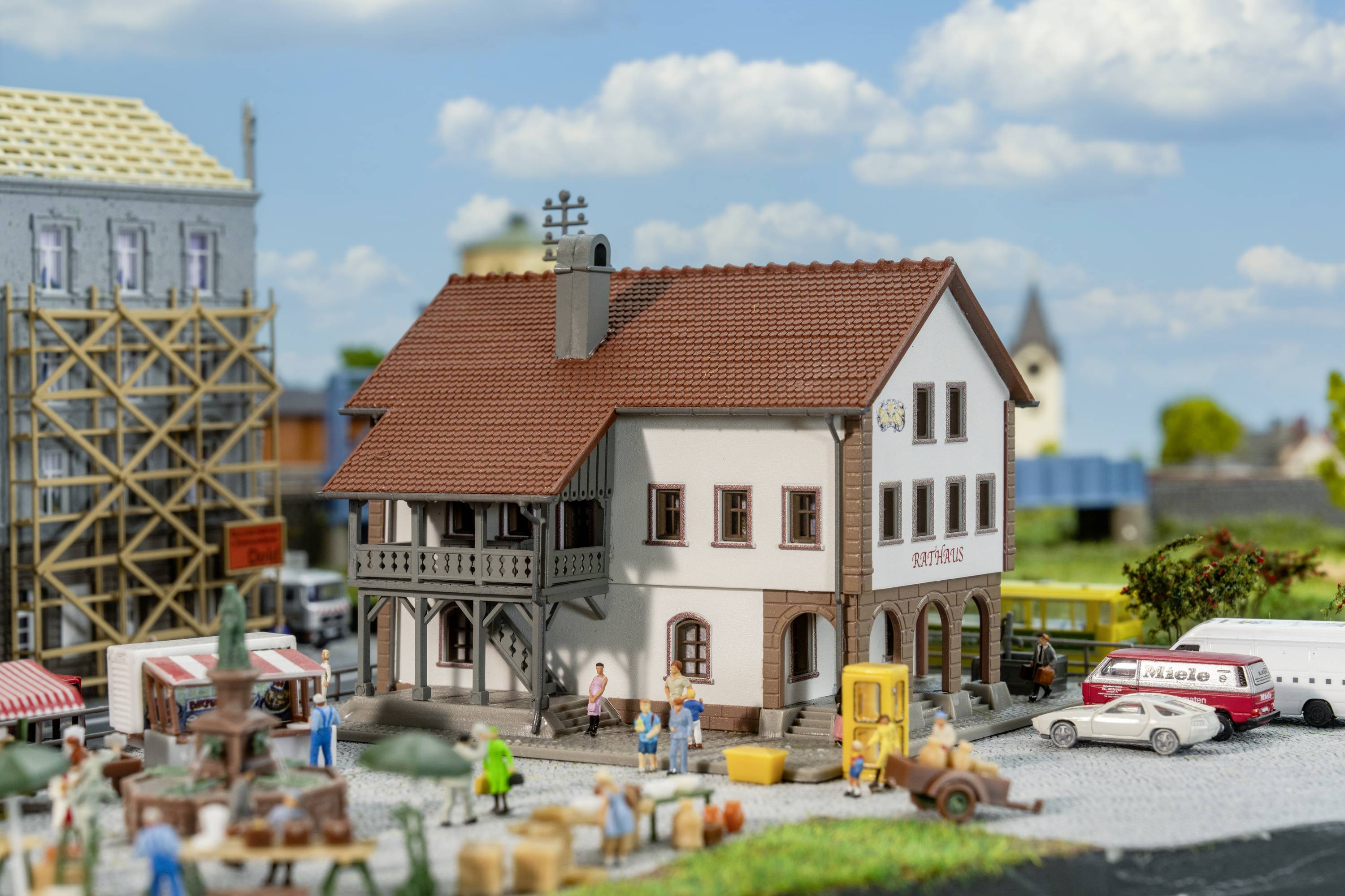 Gebäudemodell in Miniaturstadt, Fachwerkhaus mit 'Zum Adler' beschriftet. Personen und Marktstände im Vordergrund. Stimmung: lebendig.
