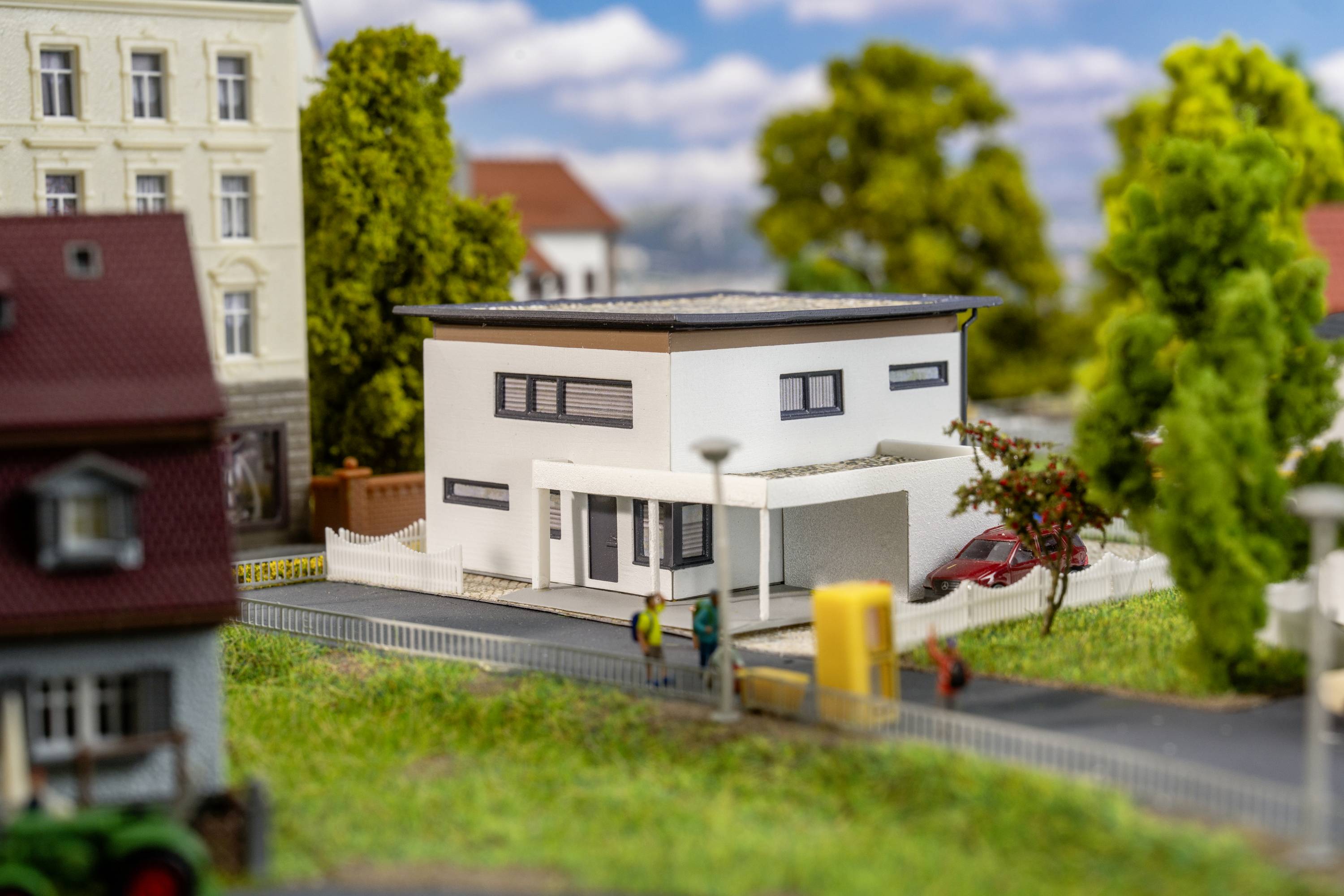 Ein modernes, weißes Einfamilienhaus mit Flachdach, umgeben von Bäumen und anderen Gebäuden. Zwei Personen stehen auf dem Gehweg.