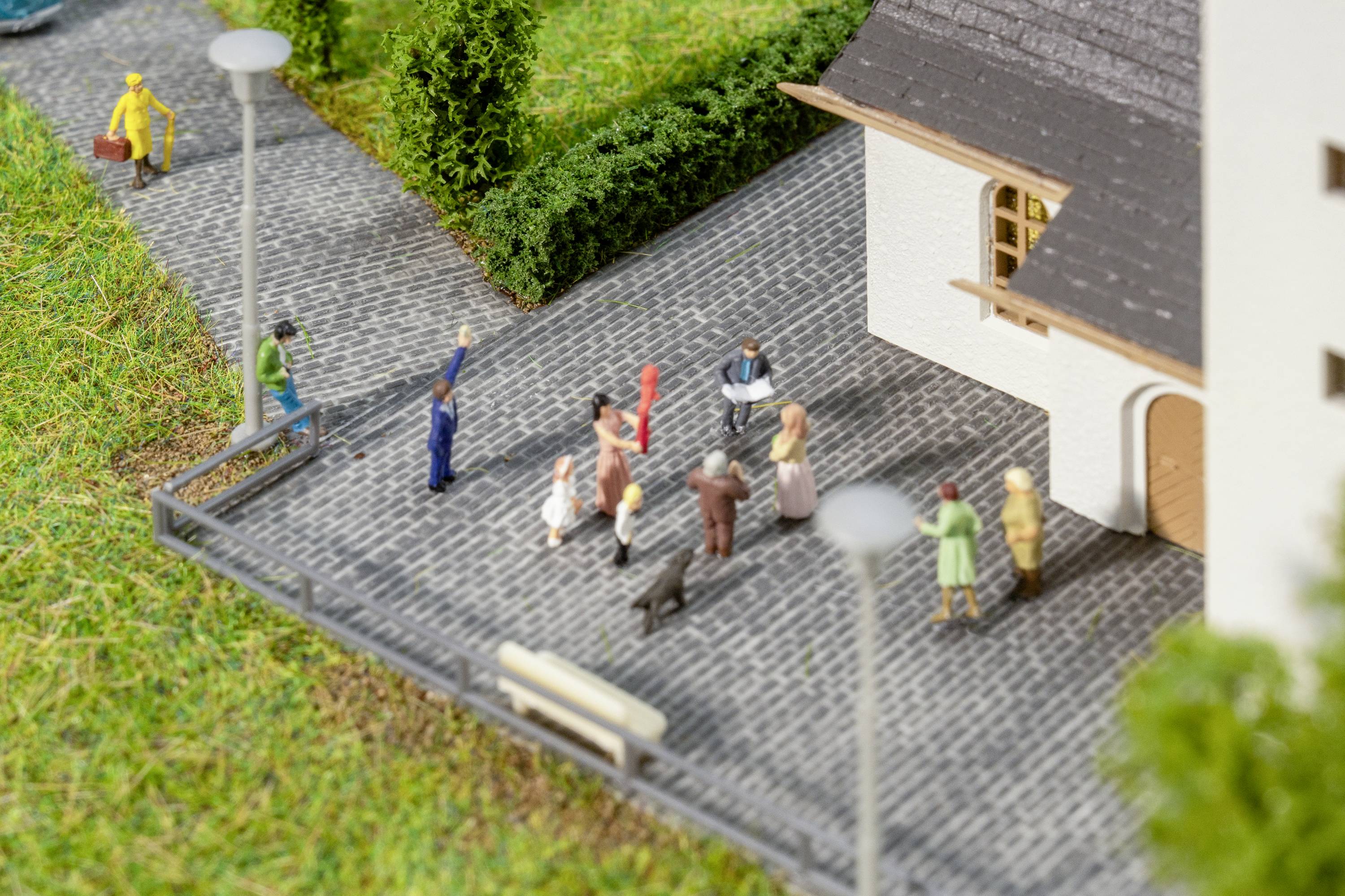 Miniaturmodell einer Kirche mit Figuren auf einem gepflasterten Weg. Menschen stehen und unterhalten sich, umgeben von Laternen.