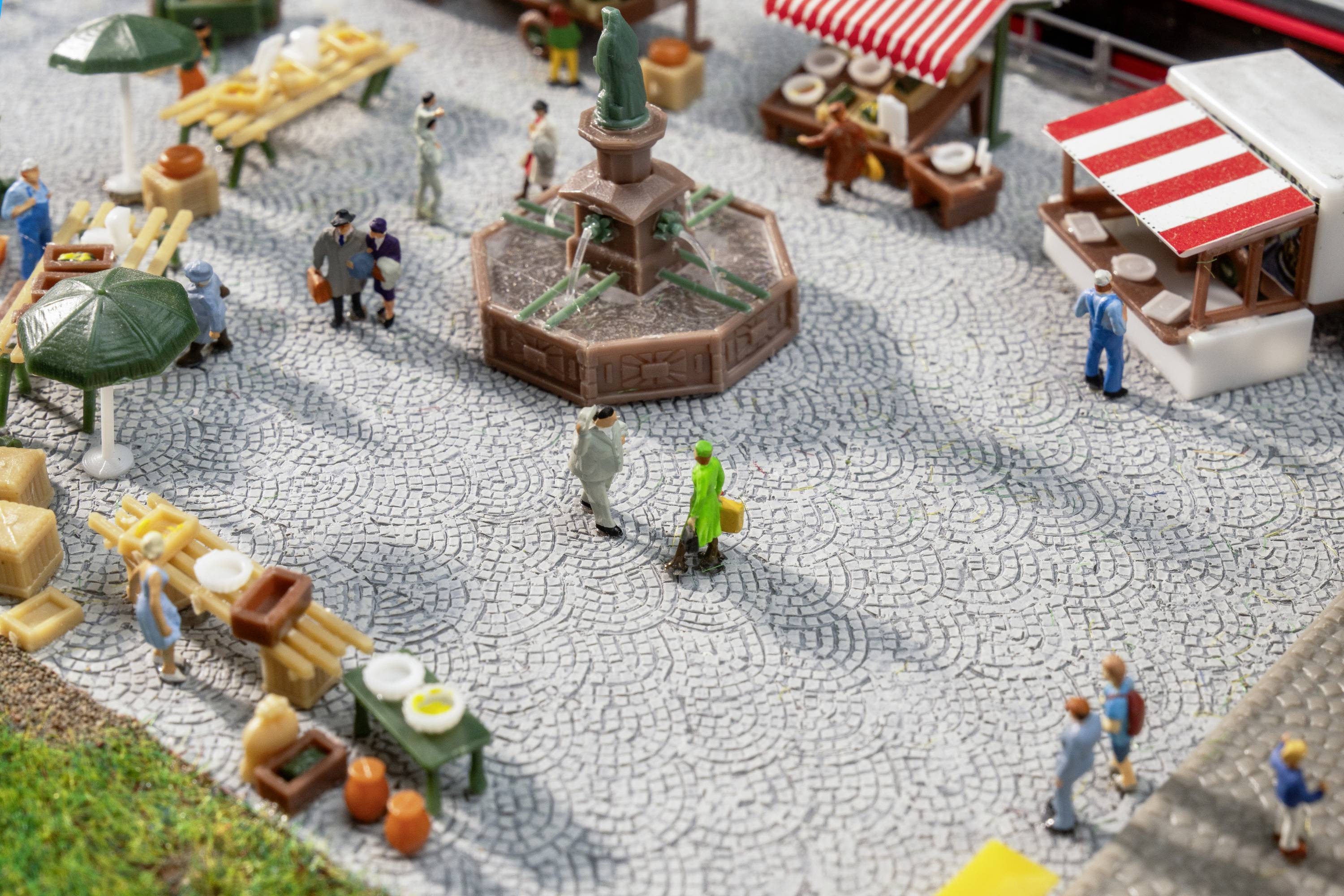 Marktszene mit Miniaturfiguren: Menschen schlendern über Platz mit Ständen und einem Brunnen in der Mitte. Lebhafte Atmosphäre.