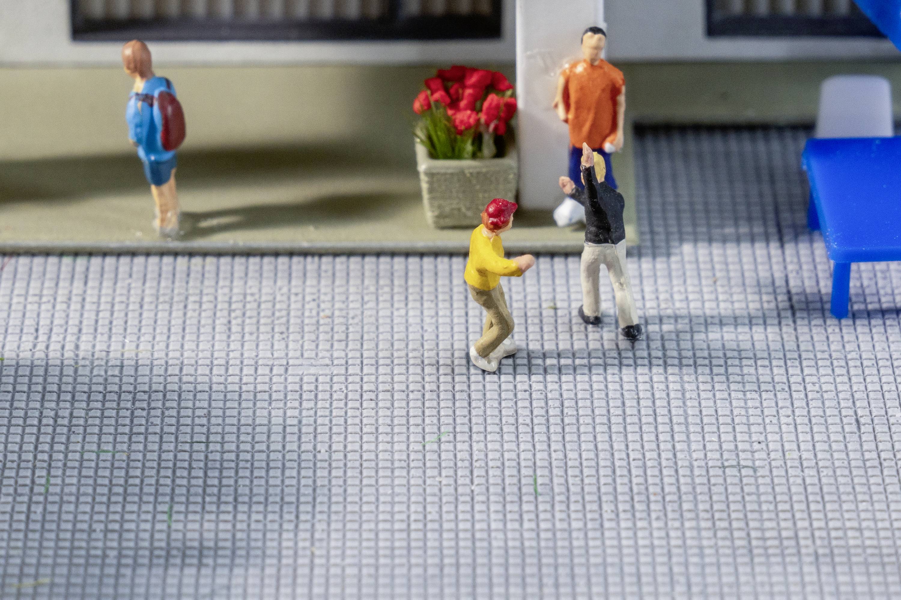 Miniaturfiguren auf einem Bürgersteig: Zwei Personen unterhalten sich, eine hält ein Blatt Papier. Blumen im Hintergrund und eine Person geht vorbei.