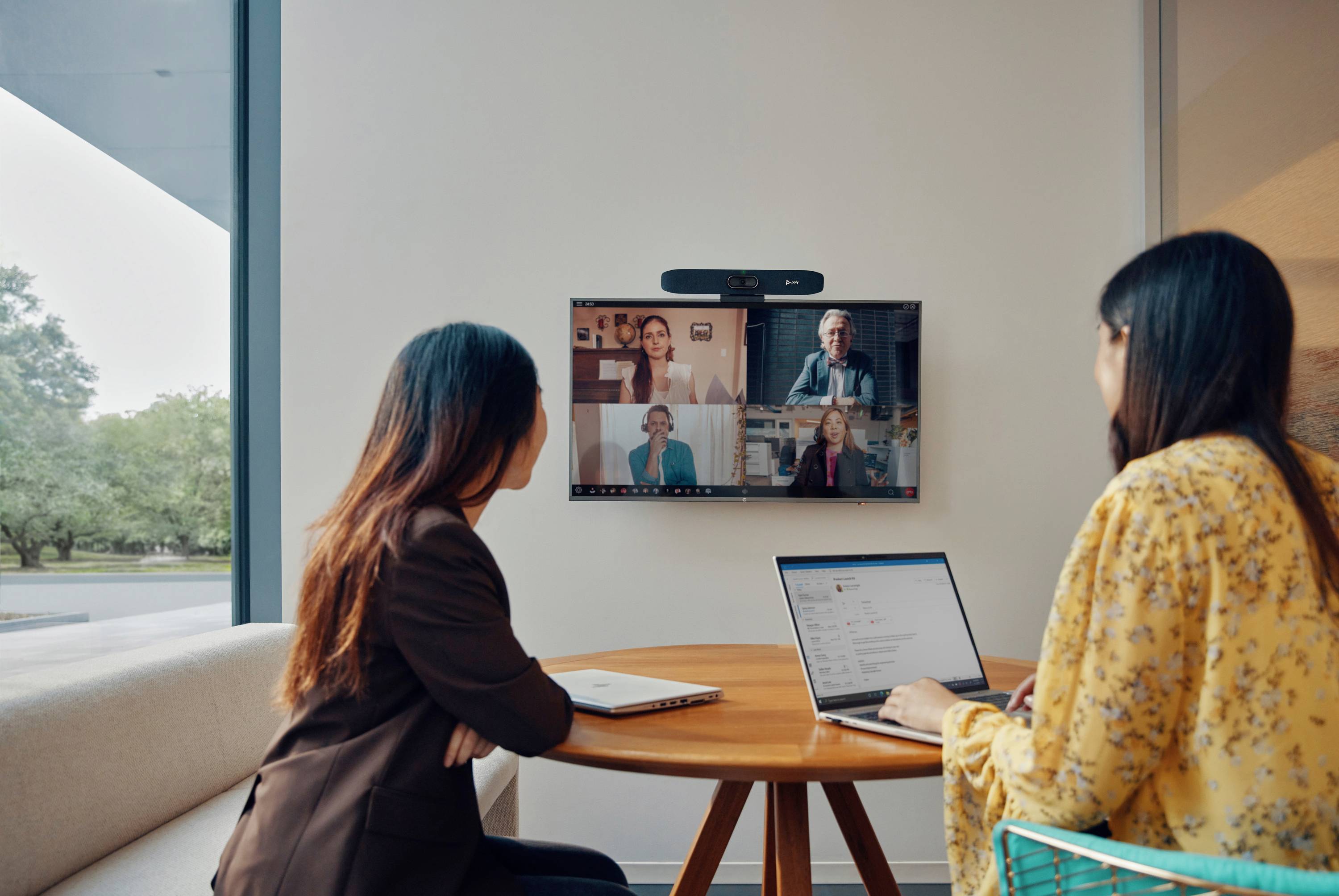 Zwei Frauen sitzen an einem Tisch und nehmen an einer Videokonferenz mit mehreren Personen auf einem Bildschirm teil.