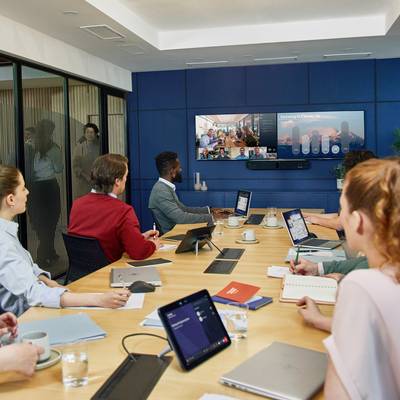 Menschen sitzen in einem Besprechungsraum mit Laptops und Notizen, während sie an einer Videokonferenz auf einem Bildschirm teilnehmen.