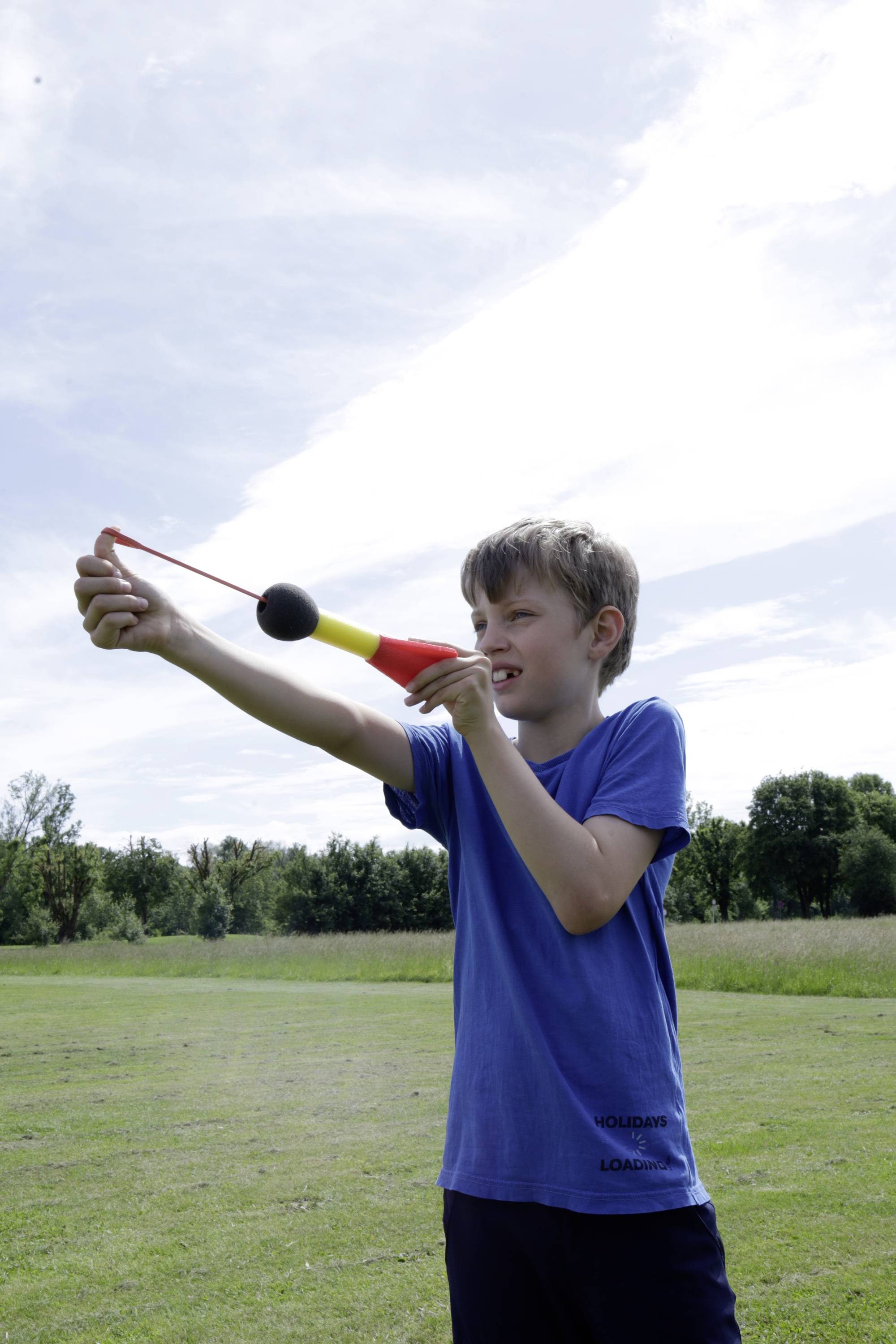 Ein Junge steht auf einer Wiese und schießt ein Ziel mit einer Schleuder. Der Himmel ist bewölkt.