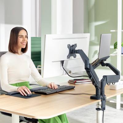 Eine Frau sitzt lächelnd an einem modernen Schreibtisch, bedient Maus und Tastatur, vor zwei Monitoren, in einem hellen Büro.