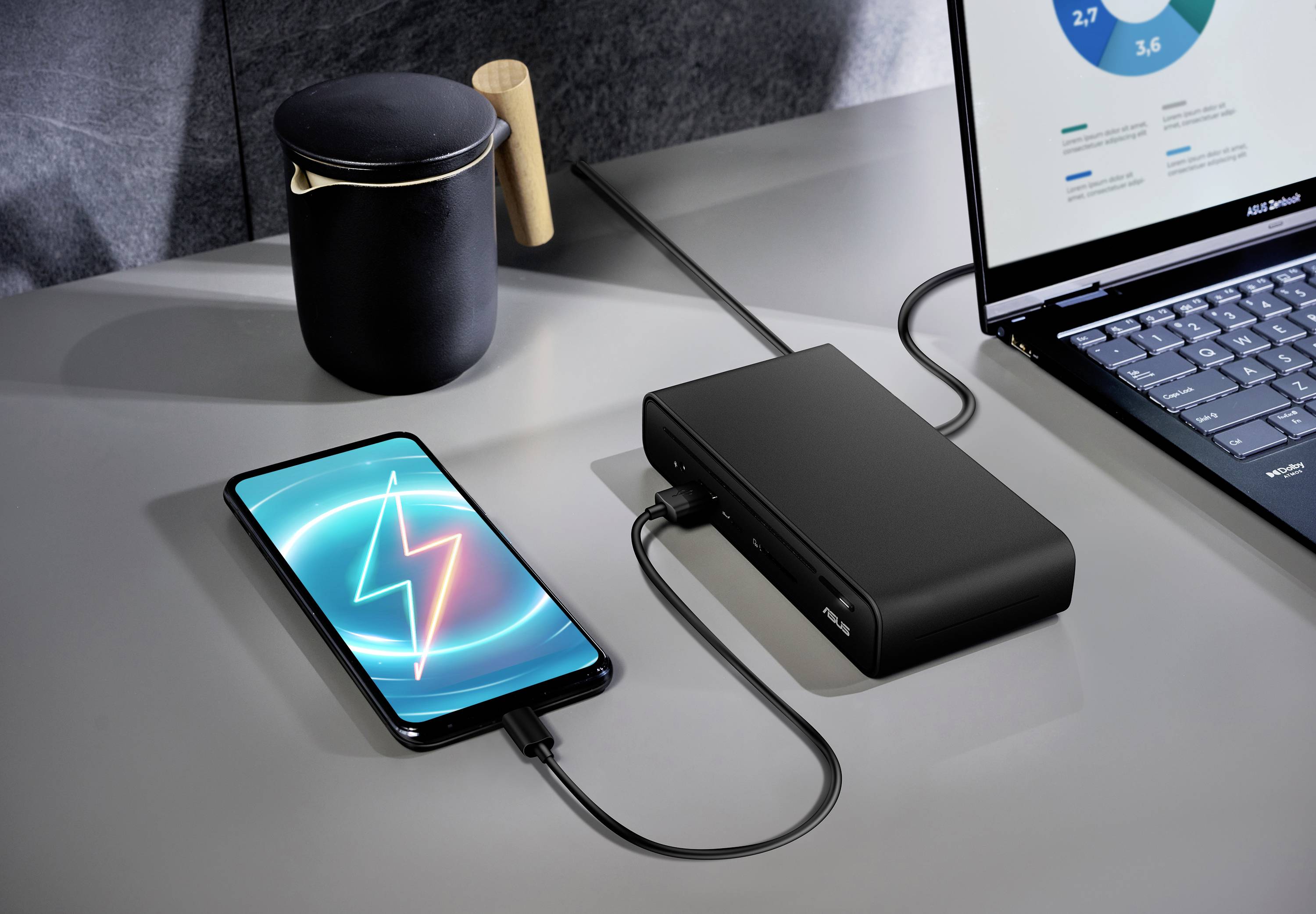 Ein Smartphone zeigt ein Ladesymbol, während es mit einer Powerbank auf einem Schreibtisch verbunden ist, neben einem Laptop und einer schwarzen Tasse.