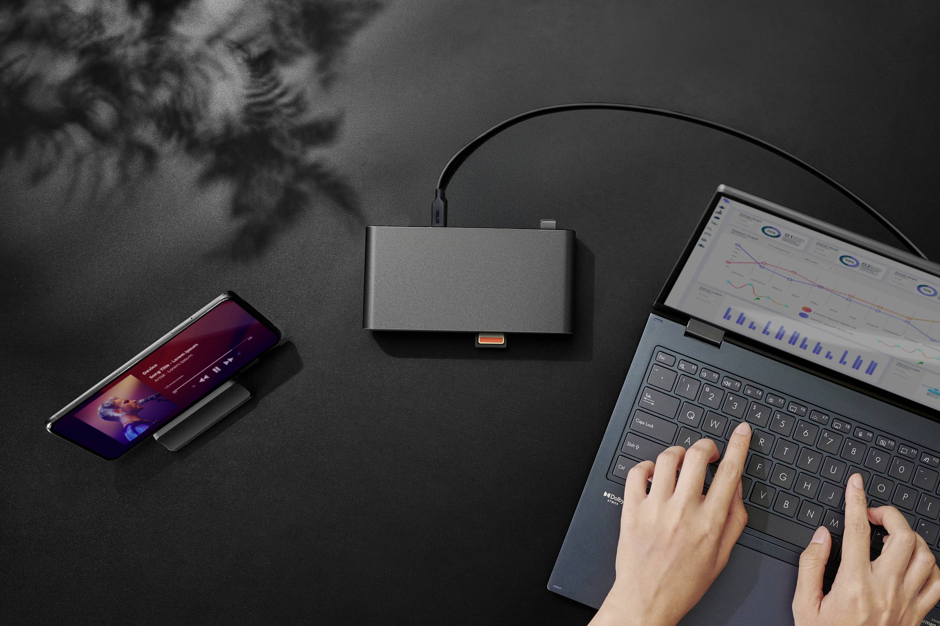 Eine Person tippt auf einem Laptop, der mit einem externen Hub verbunden ist, an dem auch ein Smartphone angeschlossen ist, das eine Musik-App anzeigt. Ein Schatten von Blättern ist im Hintergrund sichtbar.