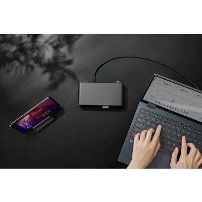 Eine Person tippt auf einem Laptop, der mit einem externen Hub verbunden ist, an dem auch ein Smartphone angeschlossen ist, das eine Musik-App anzeigt. Ein Schatten von Blättern ist im Hintergrund sichtbar.