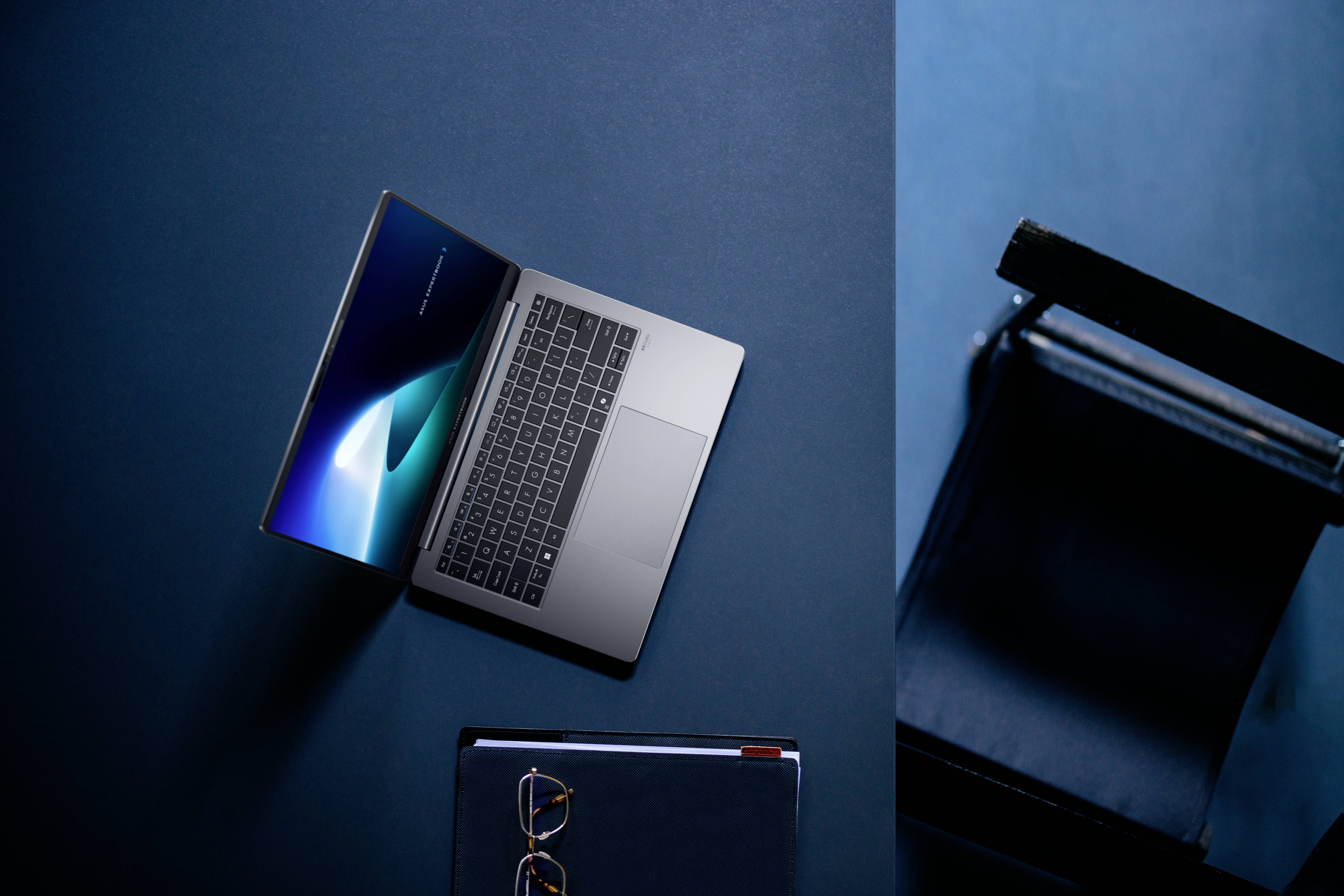 Ein Laptop mit einem leuchtenden Bildschirm ist auf einer blauen Oberfläche geöffnet, daneben liegen eine Brille und ein geschlossenes Notizbuch, und ein Stuhl steht in der Nähe.