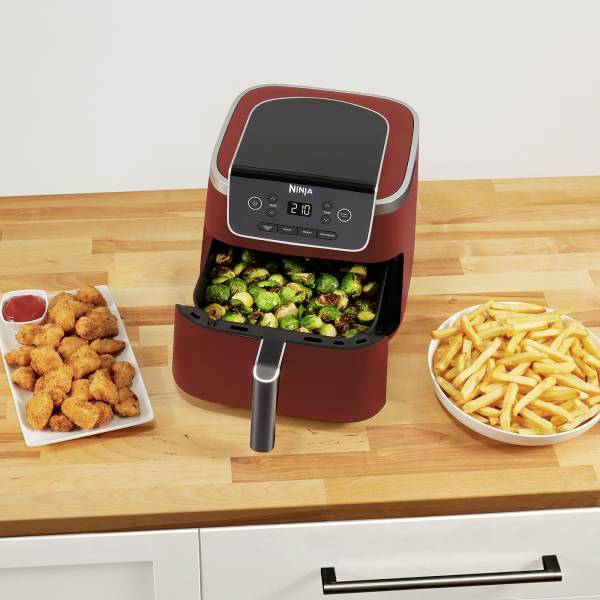 Eine Heißluftfritteuse auf einer Küchentheke mit knusprigem Gemüse darin. Daneben stehen Pommes und Hähnchen-Nuggets in Schalen.
