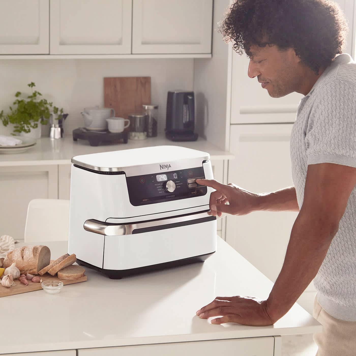 Eine Person verwendet einen kleinen, modernen Ofen in einer hellen Küche. Auf der Arbeitsplatte stehen Brote, Geschirr und Pflanzen.