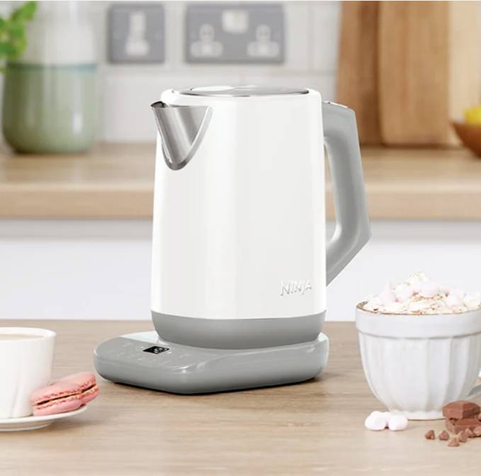 Ein weißer Wasserkocher steht auf einer Küchenarbeitsplatte neben einer Tasse mit Kakao, einer rosa Macaron und einer dekorativen Pflanze.