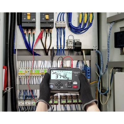 Person mit einem Multimeter in einer Elektrotafel, misst 374,6 Volt Gleichstrom, umgeben von Kabeln, Sicherungsautomaten und Steuerungen.