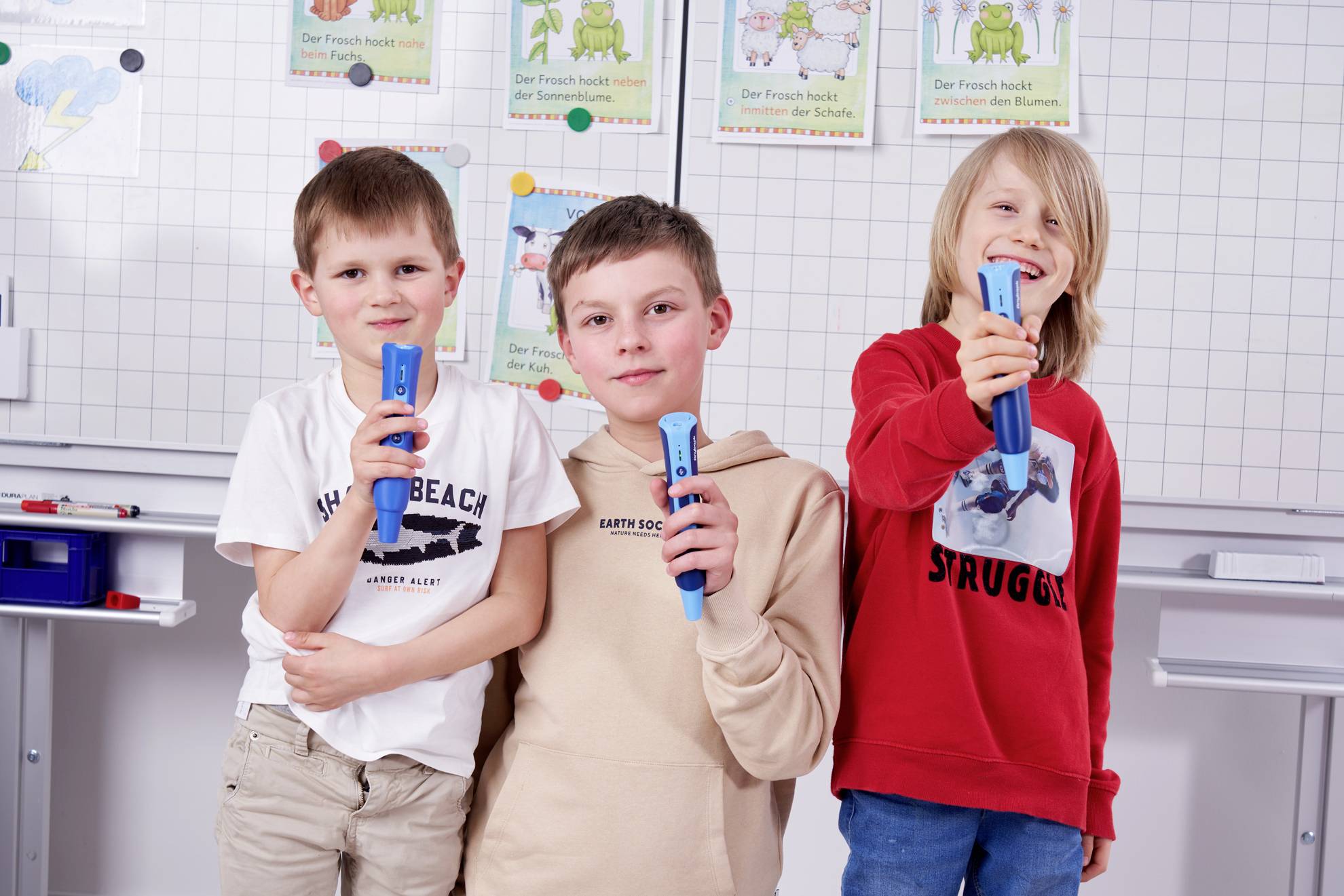 Drei Kinder posieren fröhlich mit Mikrofonen vor einer Tafel. Im Hintergrund sind kindgerechte Lernposter zu sehen.