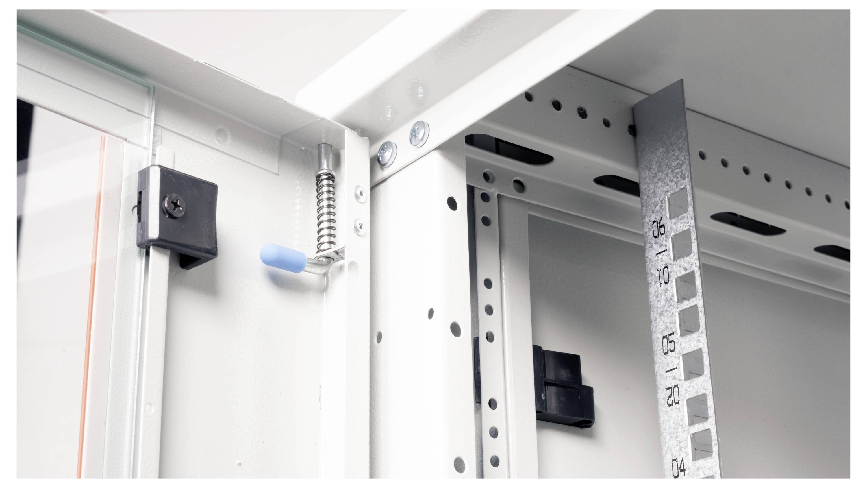 Das Bild zeigt eine Nahaufnahme des Innenraums eines metallenen Elektroschranks mit einem Federverschluss und einem vertikalen Lineal mit Maßeinteilungen.