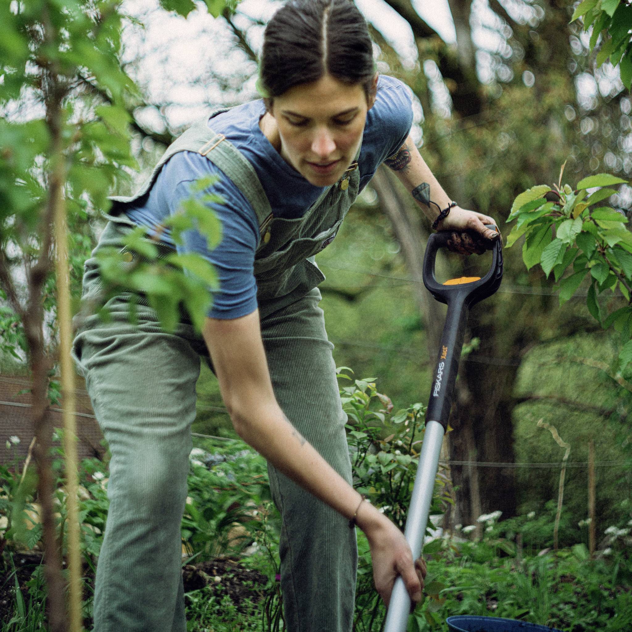 Eine Person arbeitet im Garten. Sie trägt blaue Kleidung und hält einen Spaten in der Hand. Pflanzen und Bäume sind im Hintergrund sichtbar.